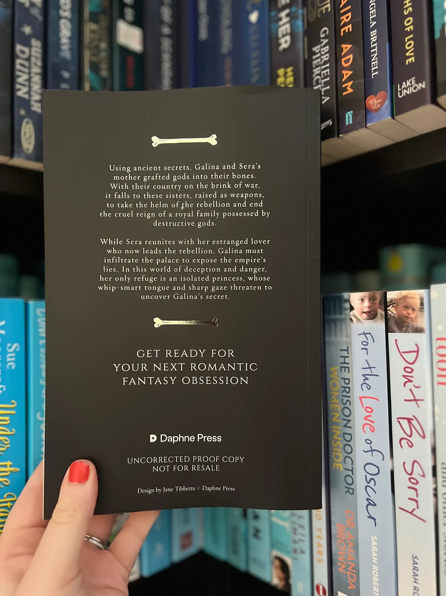 Book titled "Get Ready for Your Next Romantic Fantasy Obsession" by Daphne Press held by hand with red nails, bookshelf in background.