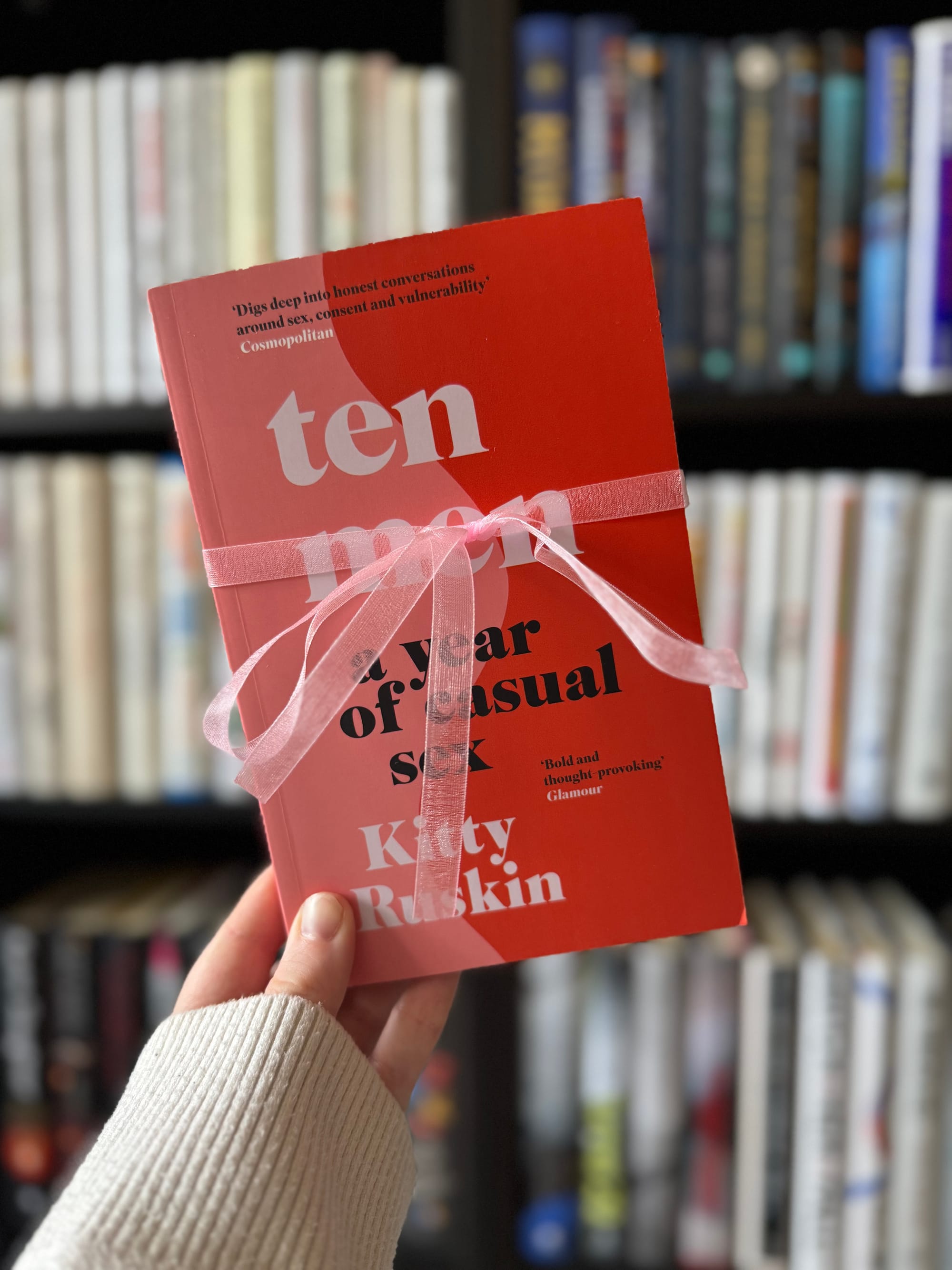 Hand holding a red book titled "Ten Men, A Year of Casual Sex" by Kitty Ruskin, wrapped in pink ribbon, with blurred bookshelves behind.