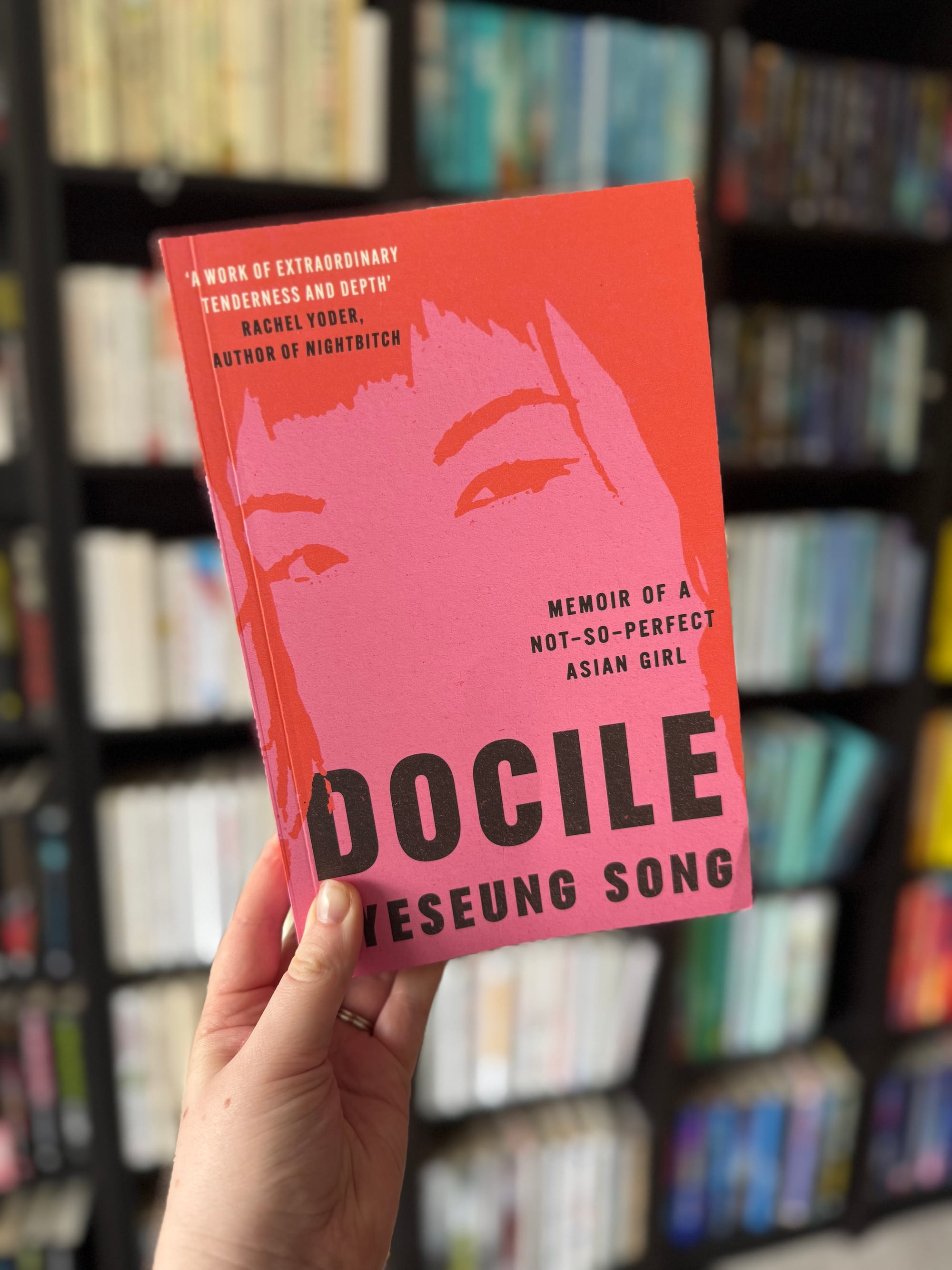Hand holds a pink book titled "DOCILE" by Heseung Song in front of a blurred bookshelf. The cover features a stylised face.