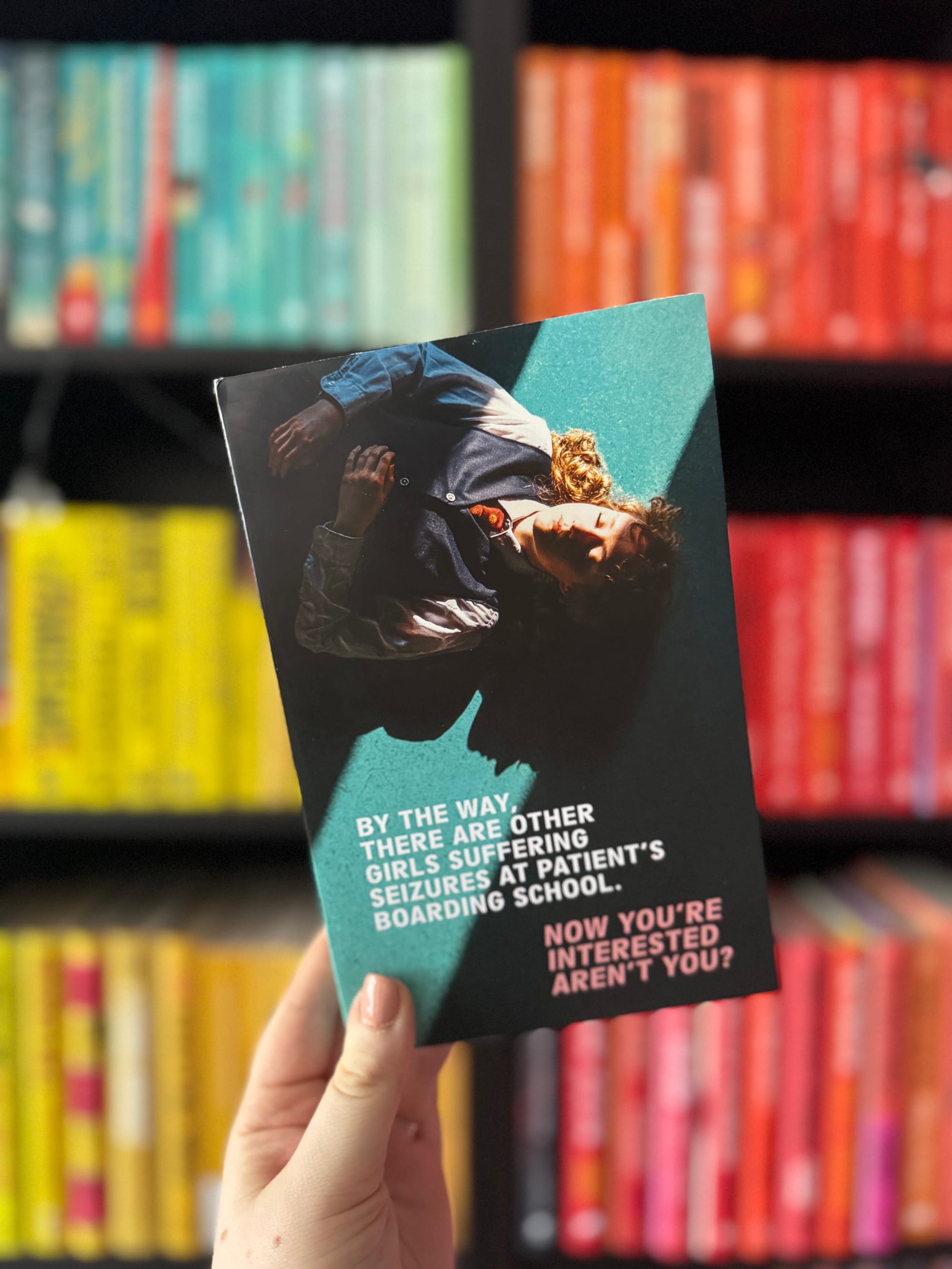 Hand holds a book with a person on cover. Text reads: "By the way, there are other girls suffering seizures at Patient's Boarding School. Now you're interested, aren't you?" Colorful shelves in background.