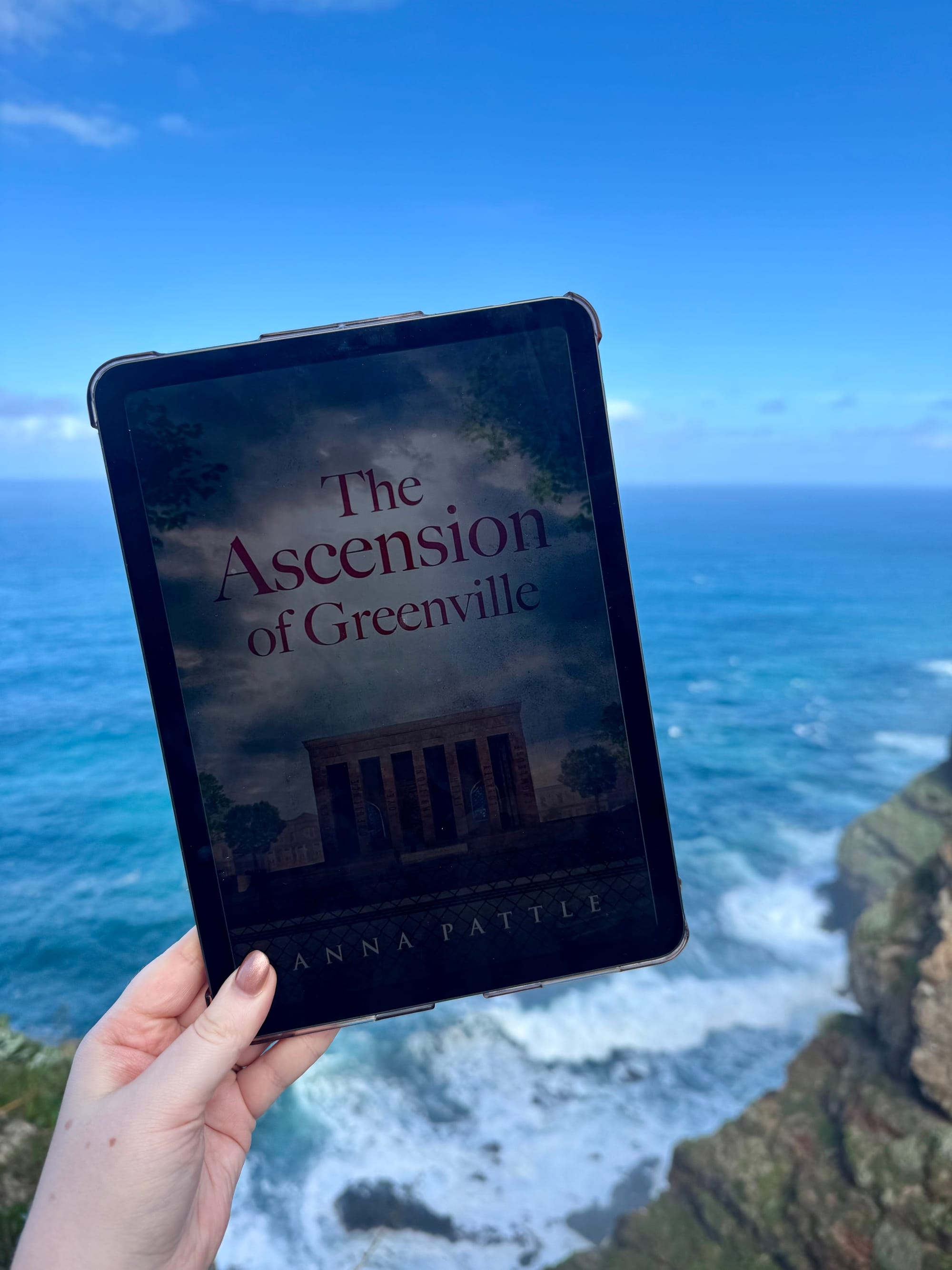 Tablet displaying "The Ascension of Greenville" is held by a hand against a cliffside backdrop with ocean waves and a clear blue sky.