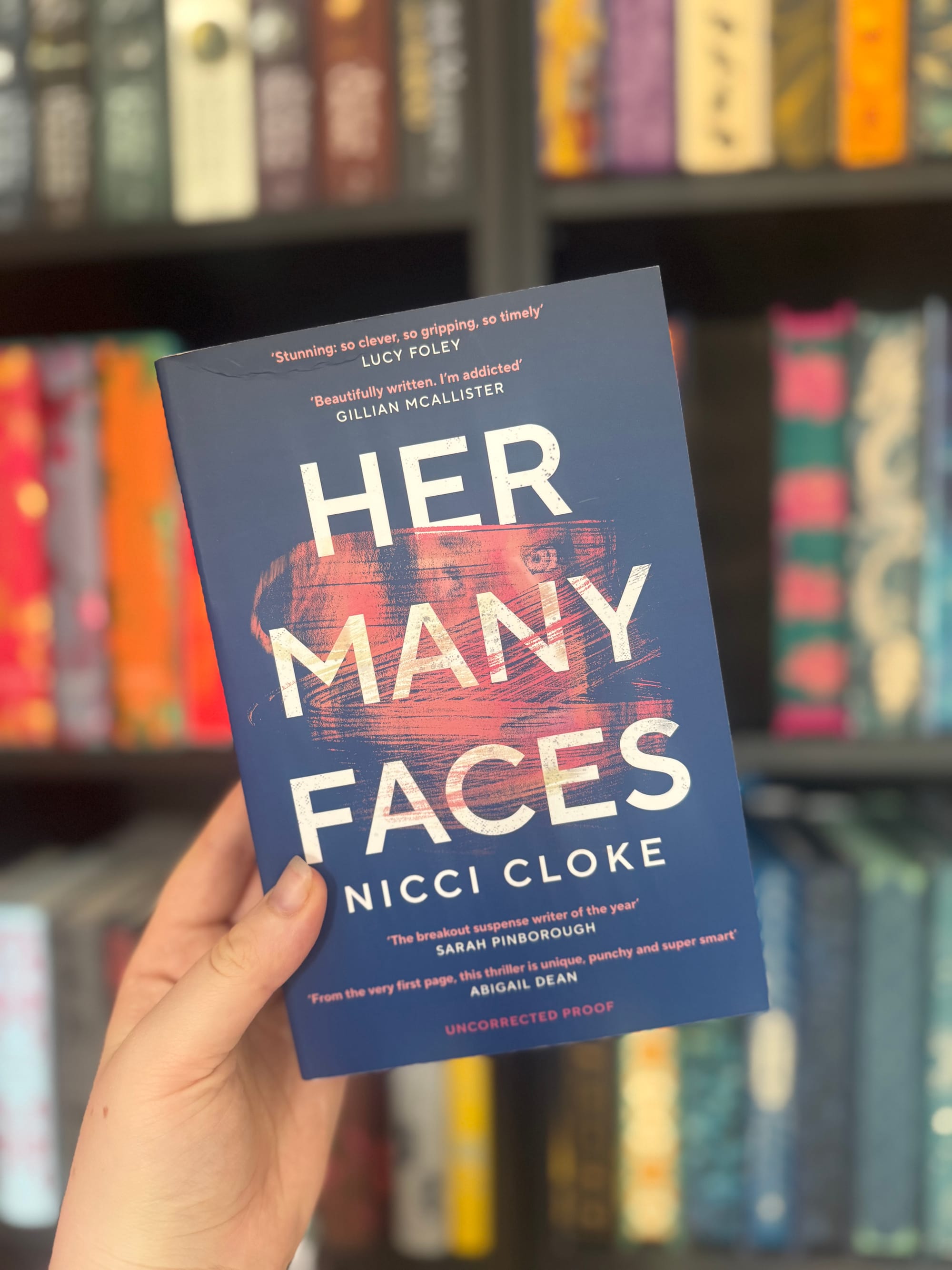 A hand holds the book "Her Many Faces" by Nicci Cloke in front of a blurred bookshelf. The book's cover is blue with pink accents.