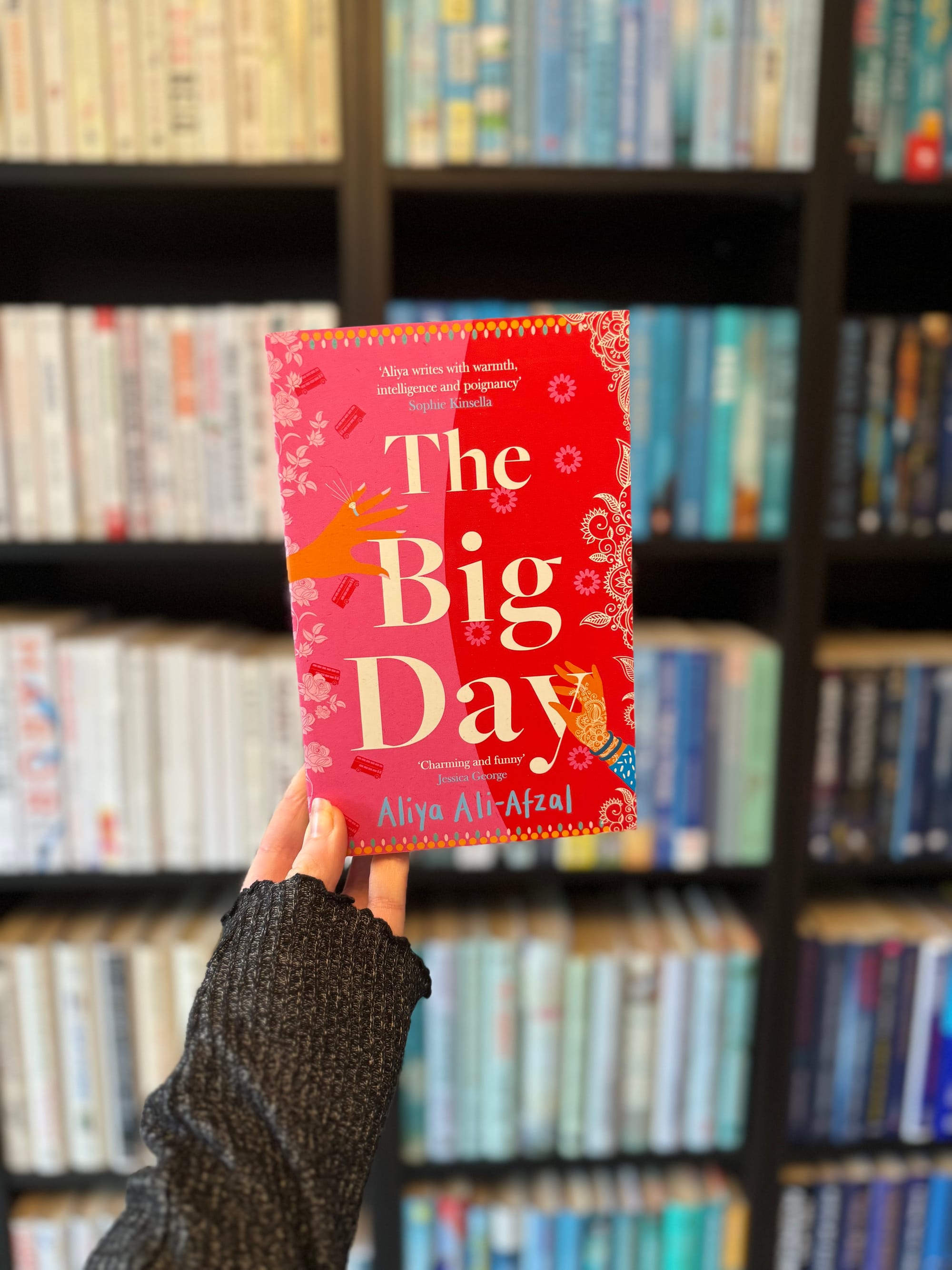 Person holding a red book titled The Big Day by Aliya Ali-Afzal in front of a blurred bookshelf. The book features floral patterns.