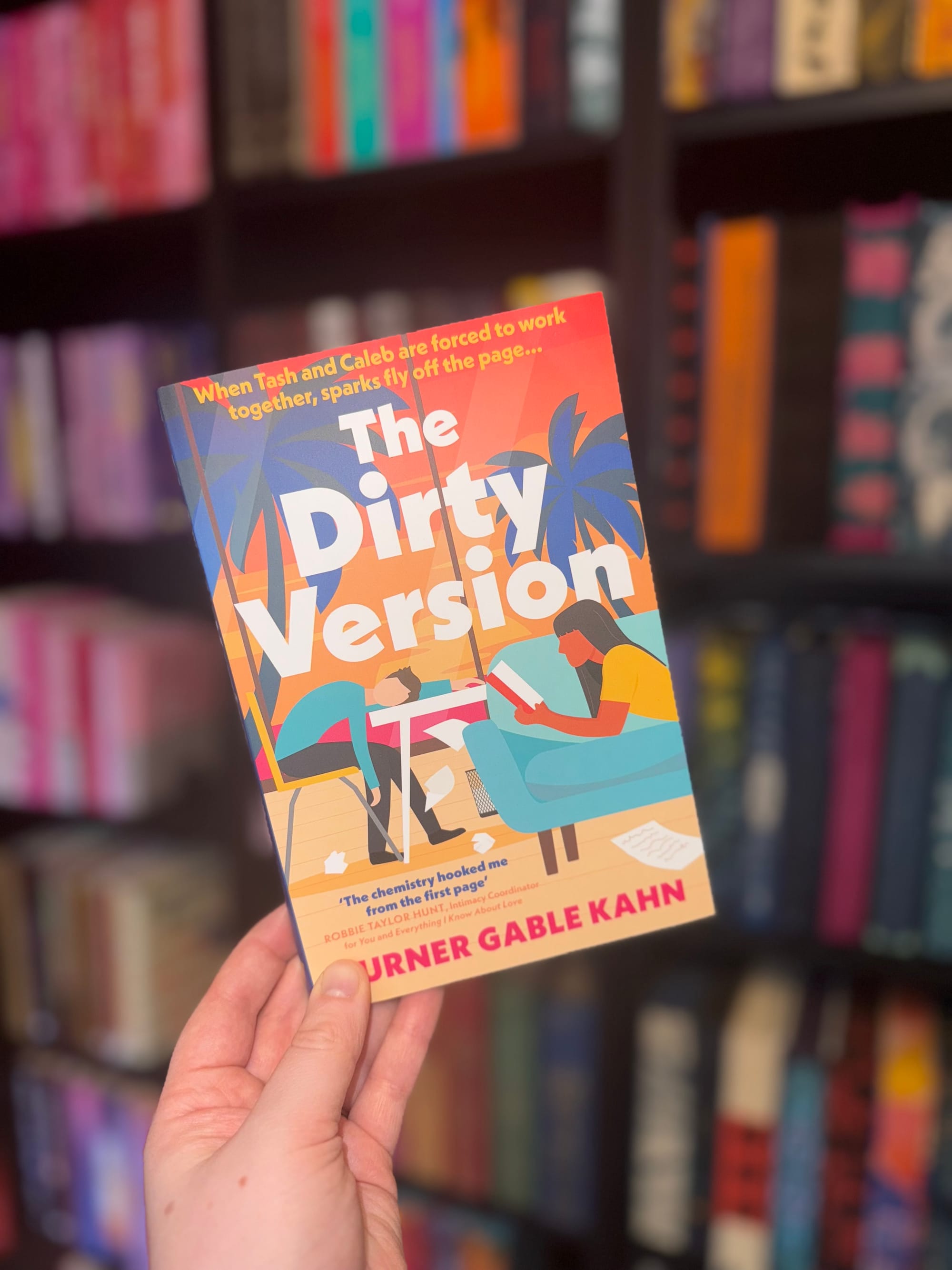 Hand holding "The Dirty Version" book with colorful cover art of two people working. Text: "When Tash and Caleb...," bookshelf backdrop.