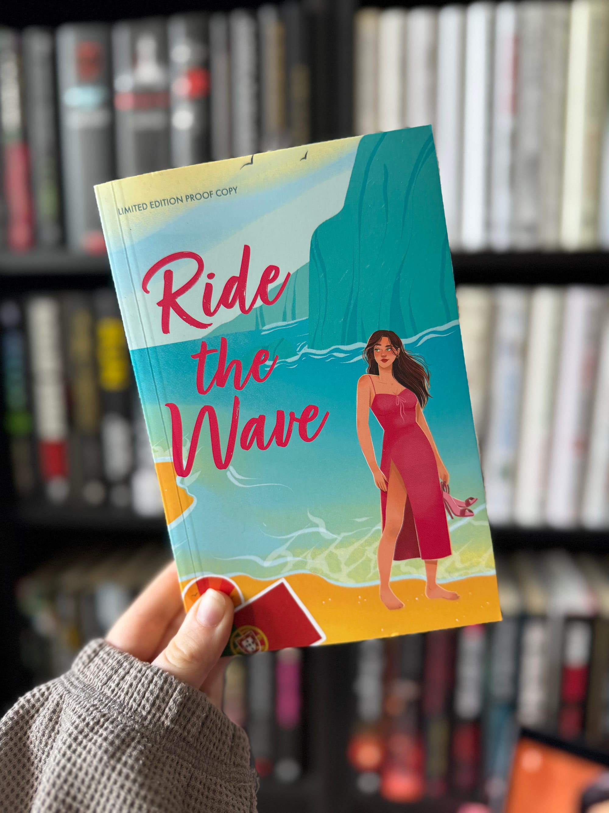 Hand holds a colorful book titled "Ride the Wave" with a woman in a red dress on the cover. Bookshelf blurred in the background. Cozy mood.