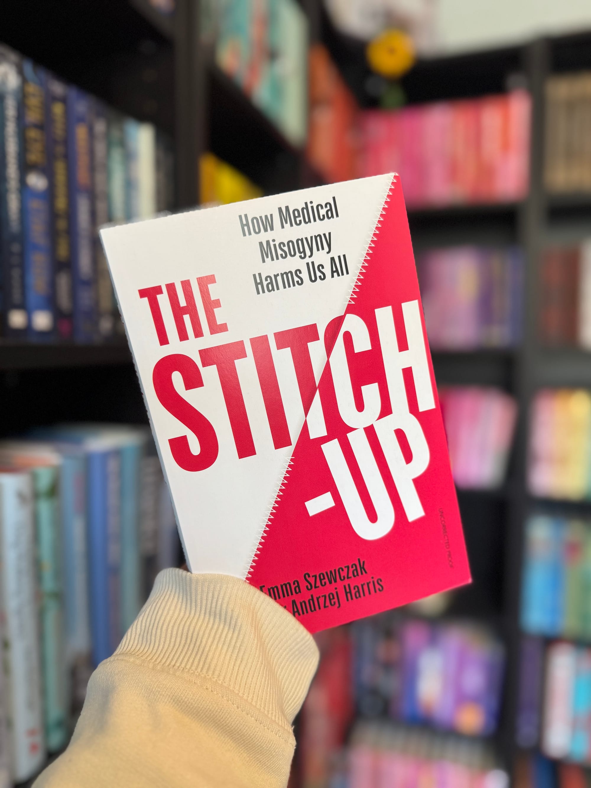 Person holds a book titled "The Stitch-Up: How Medical Misogyny Harms Us All" with red and white cover in a colorful bookstore setting.