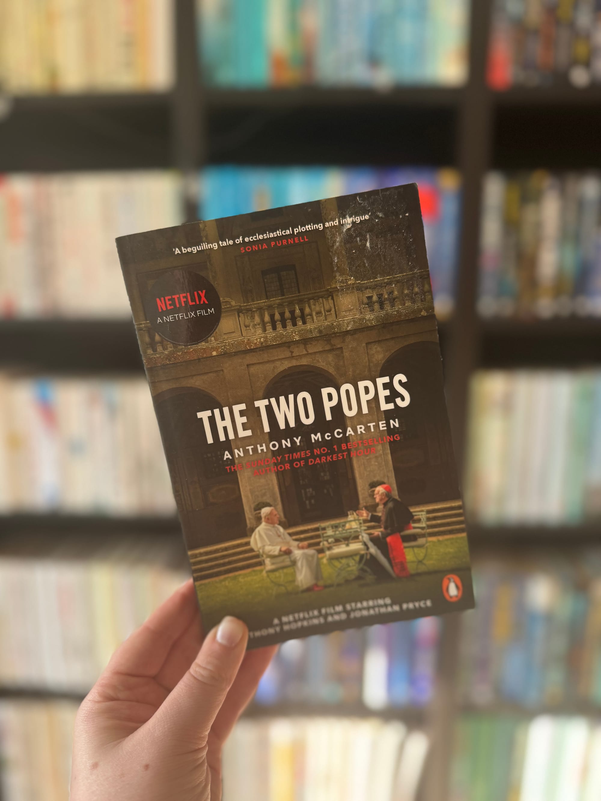 A hand holds "The Two Popes" book by Anthony McCarten. Netflix branding is visible. Blurry bookshelf background with colorful spines.