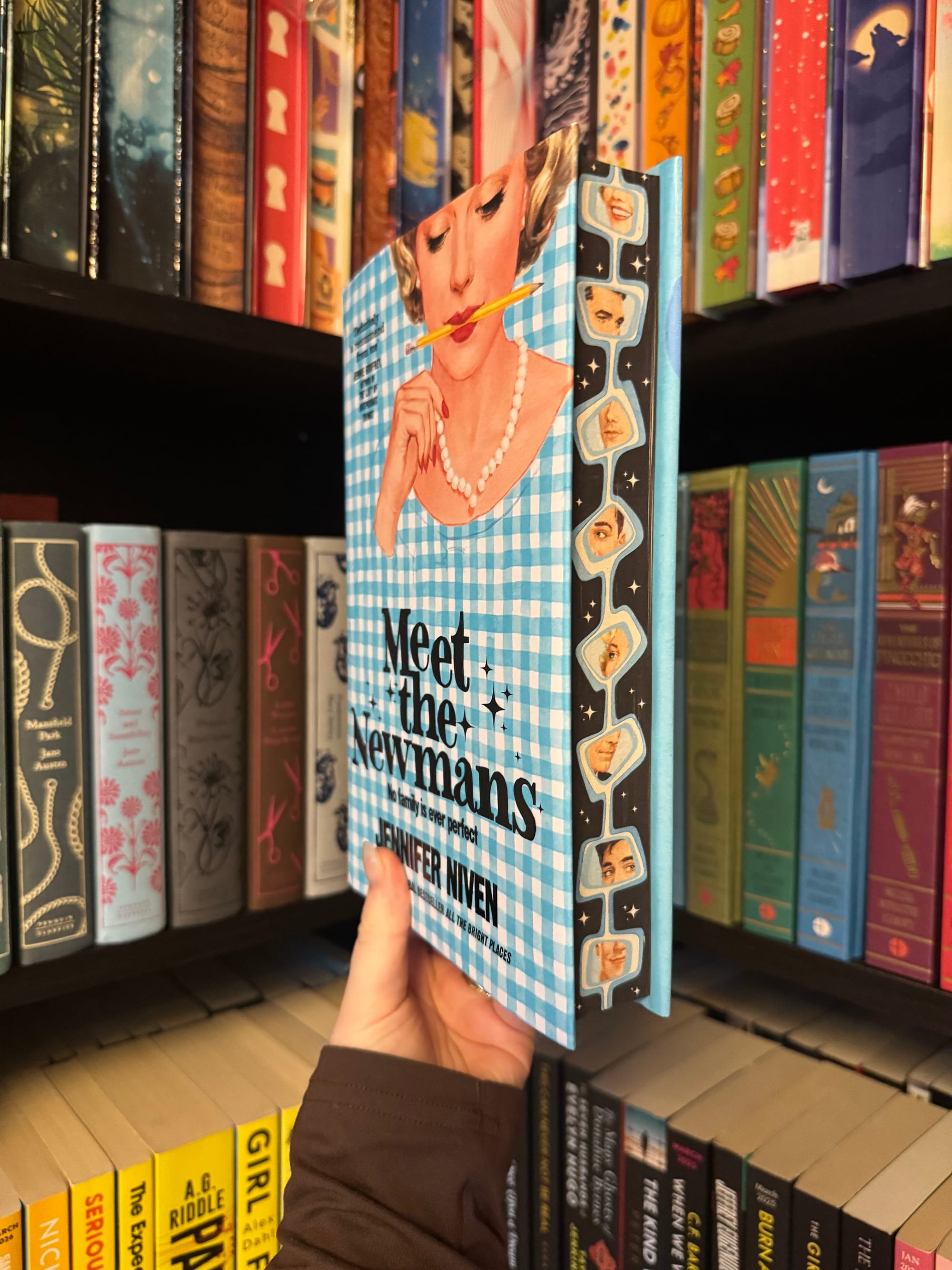 A hand holds a hardback copy of Meet the Newmans by Jennifer Niven in front of a bookshelf. The cover features a retro illustration of a woman with a pencil at her lips on a blue gingham background, with decorative illustrated endpapers visible along the book’s edge.