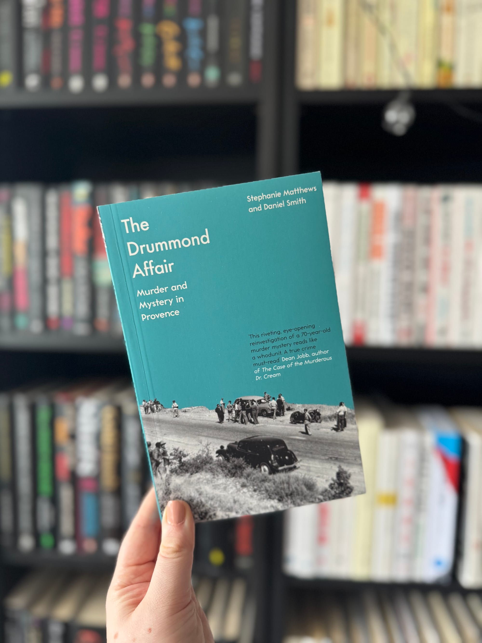 A hand holds a book titled "The Drummond Affair" against a bookshelf. The cover is teal with a vintage photo. Books are blurred in the background.