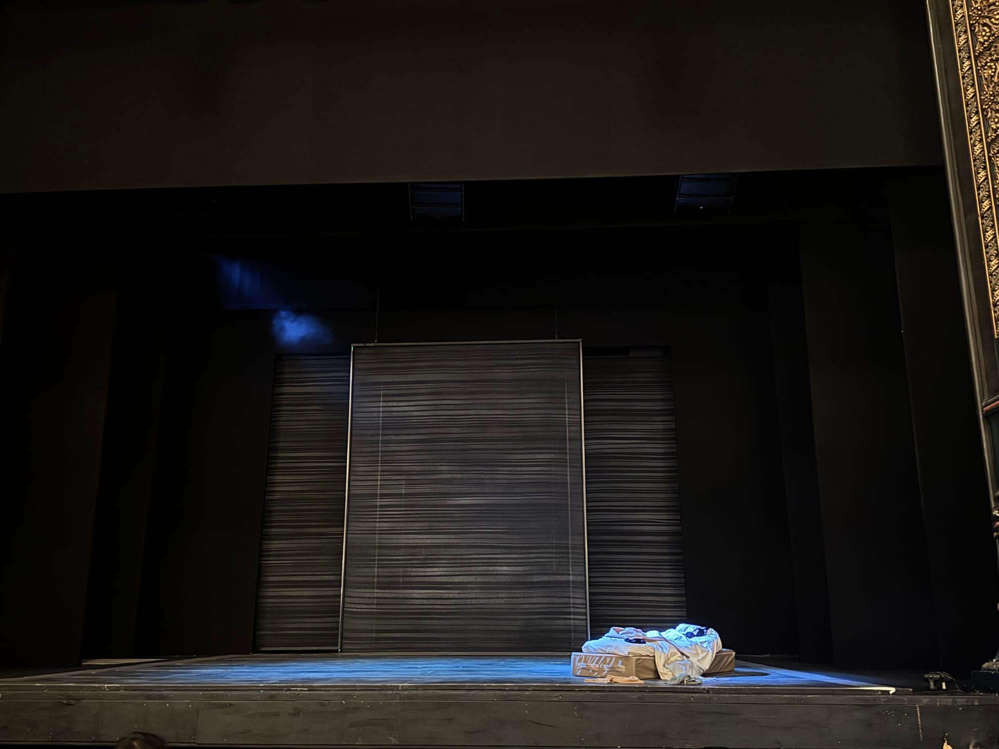 View of the stage set for The Girl on the Train at Leeds Grand Theatre. The stage is minimalist, featuring a large dark panel backdrop and a spotlight on a pile of crumpled bedding and bags on the floor. A wisp of stage smoke drifts from the left, creating a tense atmosphere.