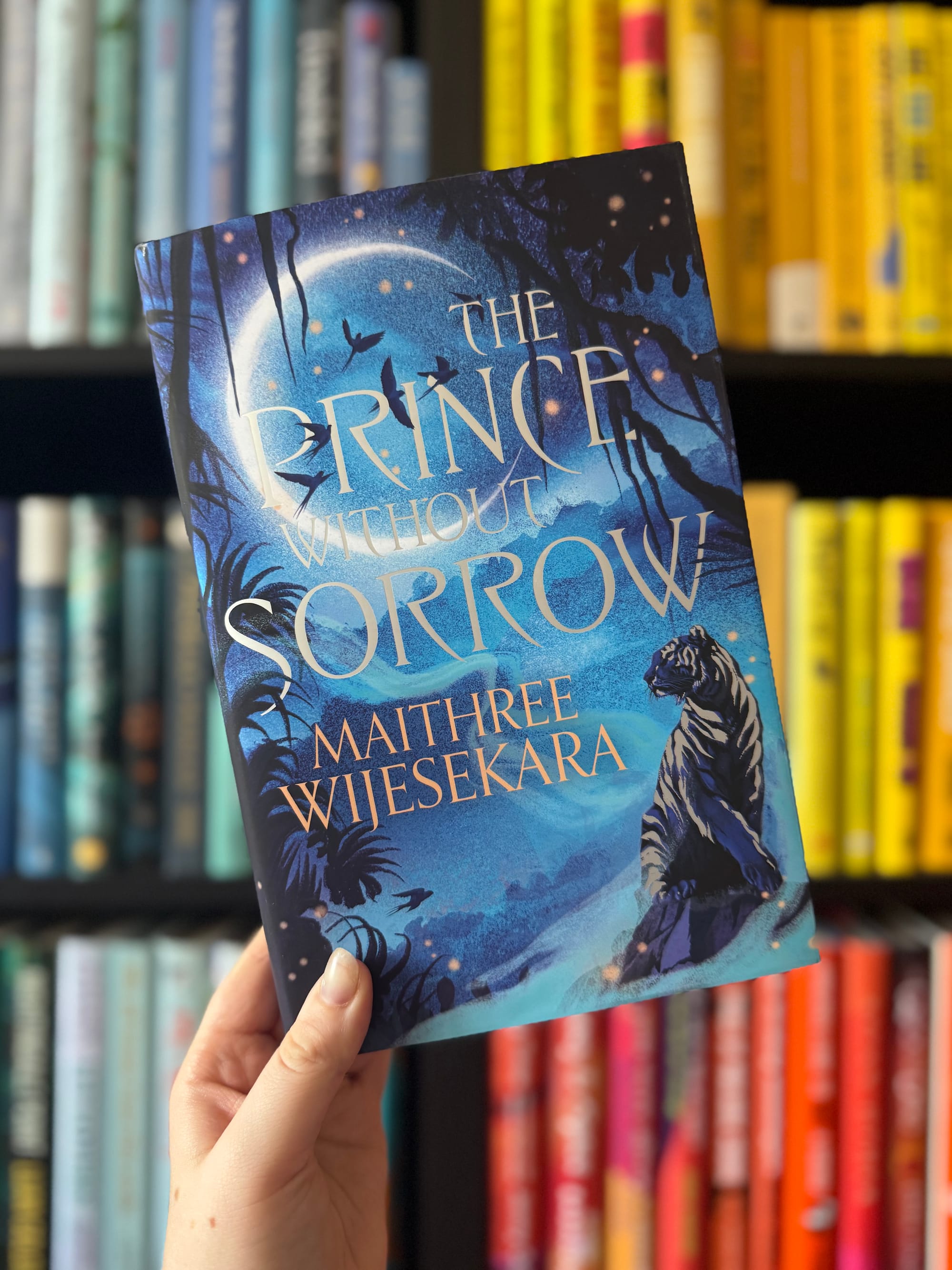 A hand holds a book titled "The Prince Without Sorrow" by Maithree Wijesekara. Cover features blue tones, moonlit scene with a tiger. Bookshelf behind.