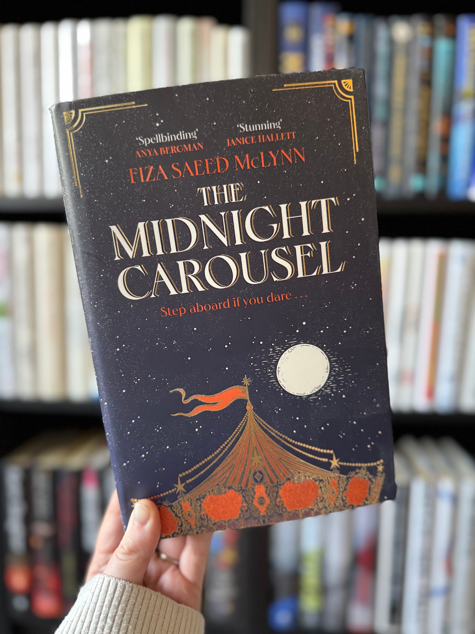 Person holding a book titled "The Midnight Carousel" by Fiza Saeed McLynn. Navy cover with a circus tent under a full moon. Bookshelf background.