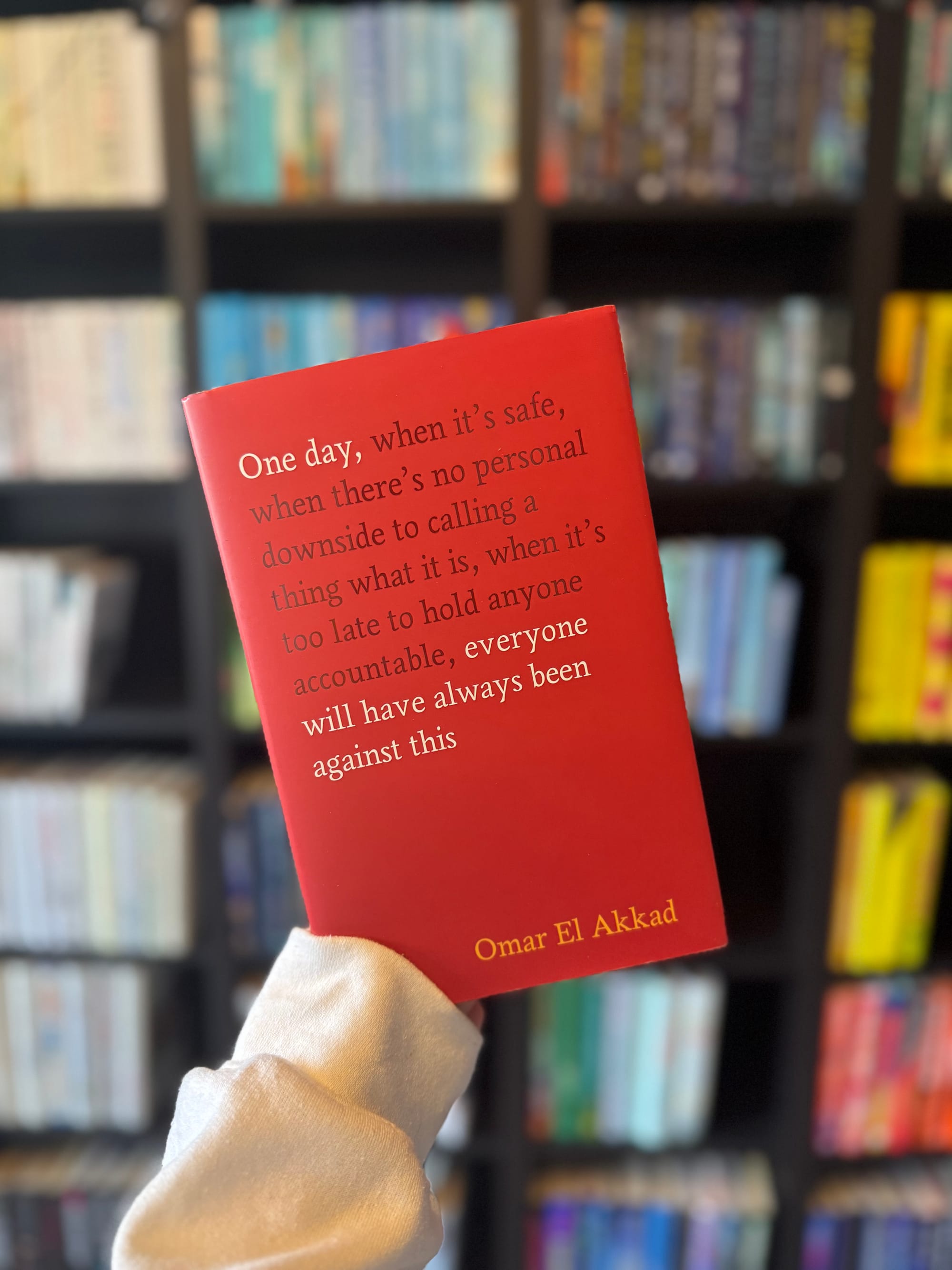 Person holding a red book with a quote by Omar El Akkad against a blurred bookshelf background, creating a thoughtful mood.