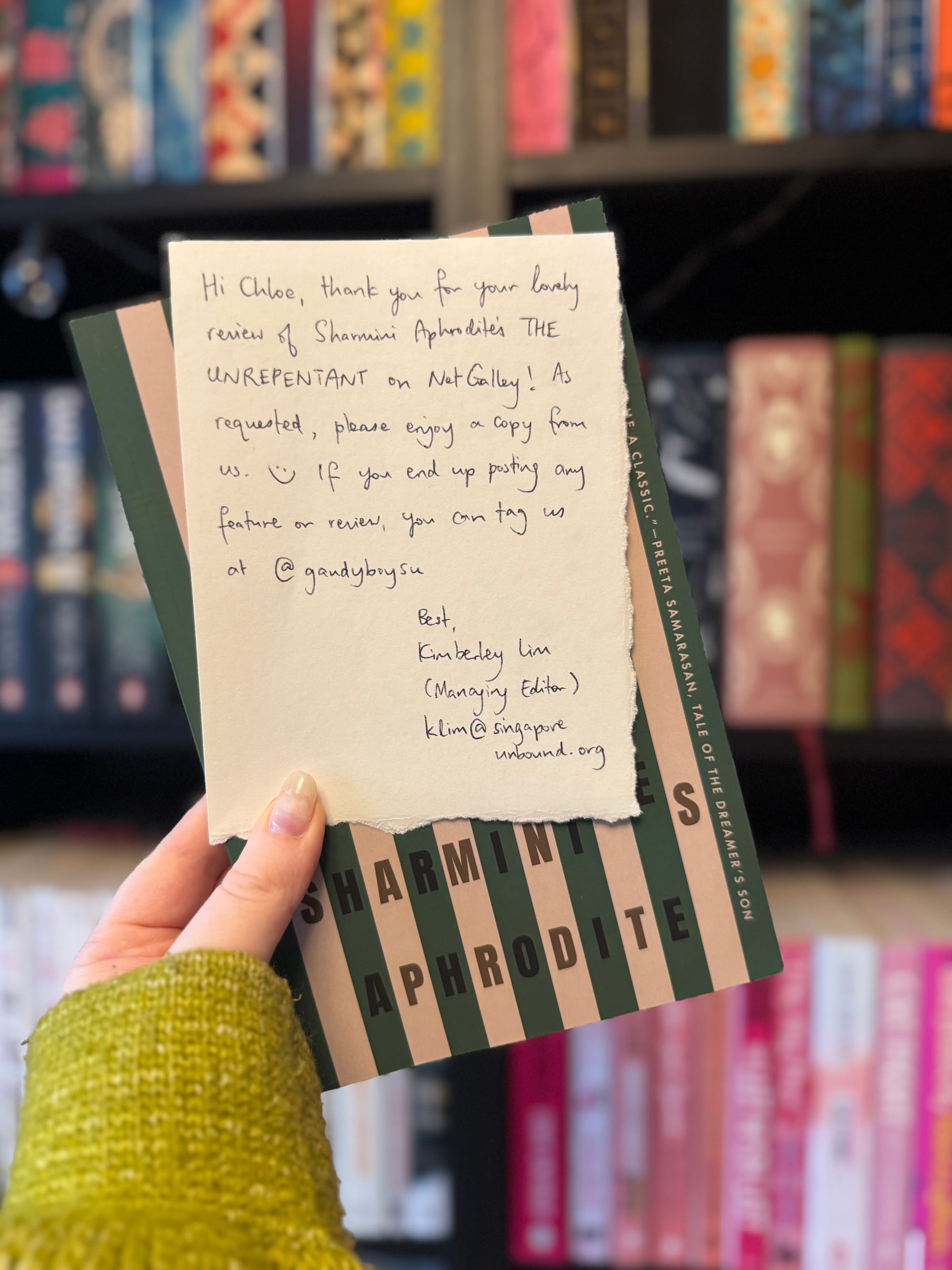 Hand holding a book with green stripes. A note on top thanks Chloe for her review and offers a copy, with colorful bookshelves in the background.