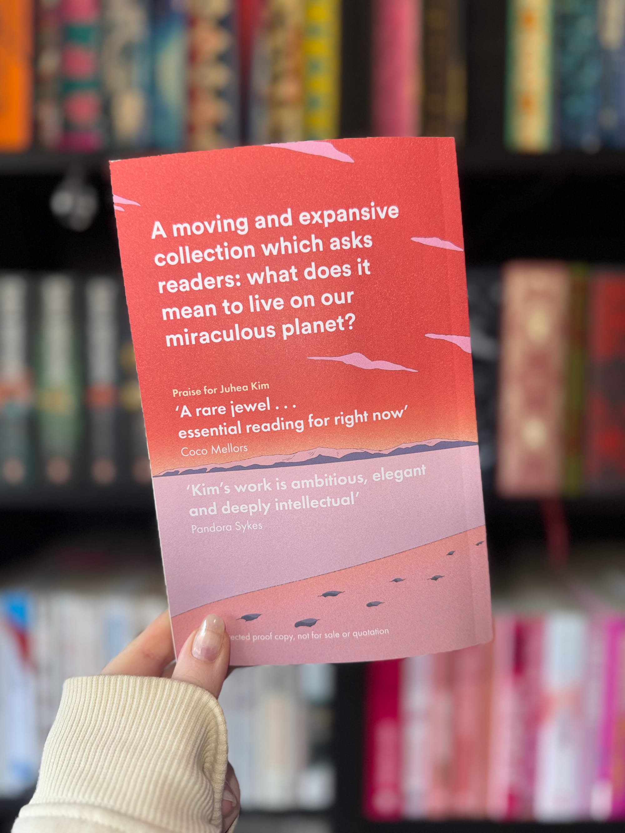 Hand holding a book with a red and pink cover. Text reads: "A moving and expansive collection... to live on our miraculous planet?" Background blurred with books.