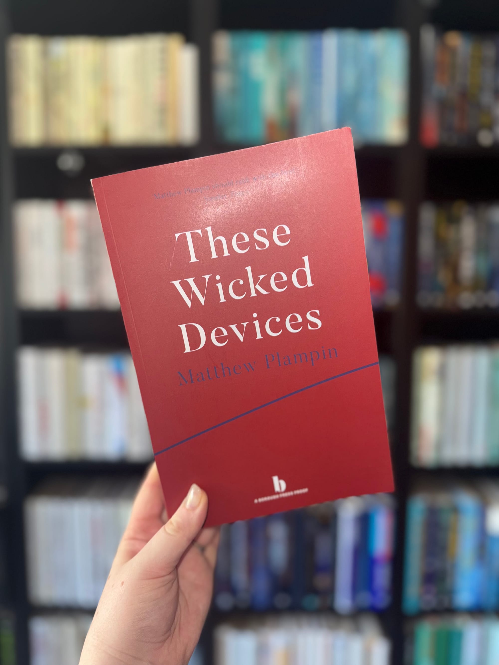 Hand holding a red book titled "These Wicked Devices" by Matthew Plampin. Background features blurred bookshelves filled with books.