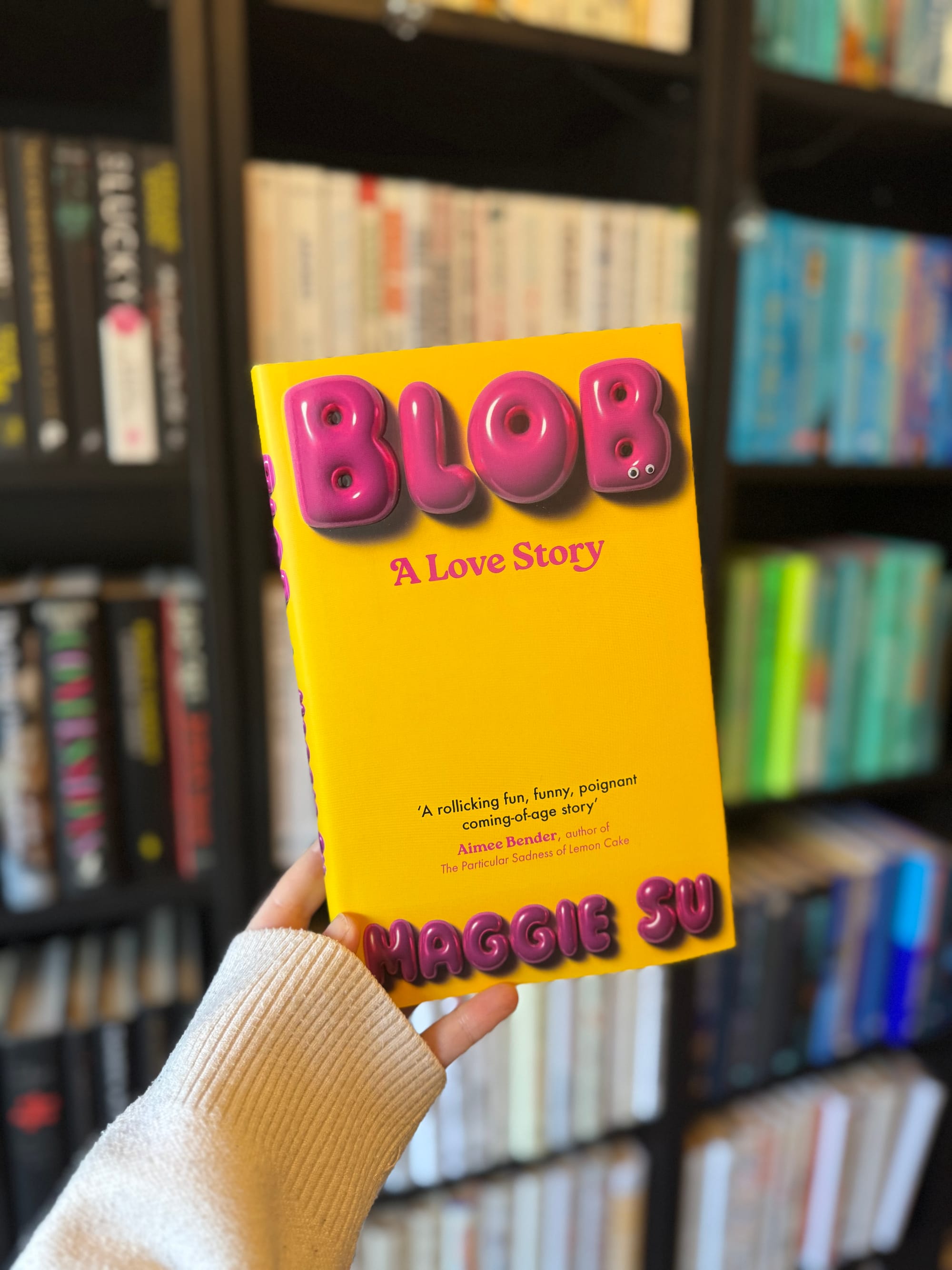 A hand holds a yellow book titled "Blob: A Love Story" against a blurry bookshelf. The cover features bold, puffy pink letters.