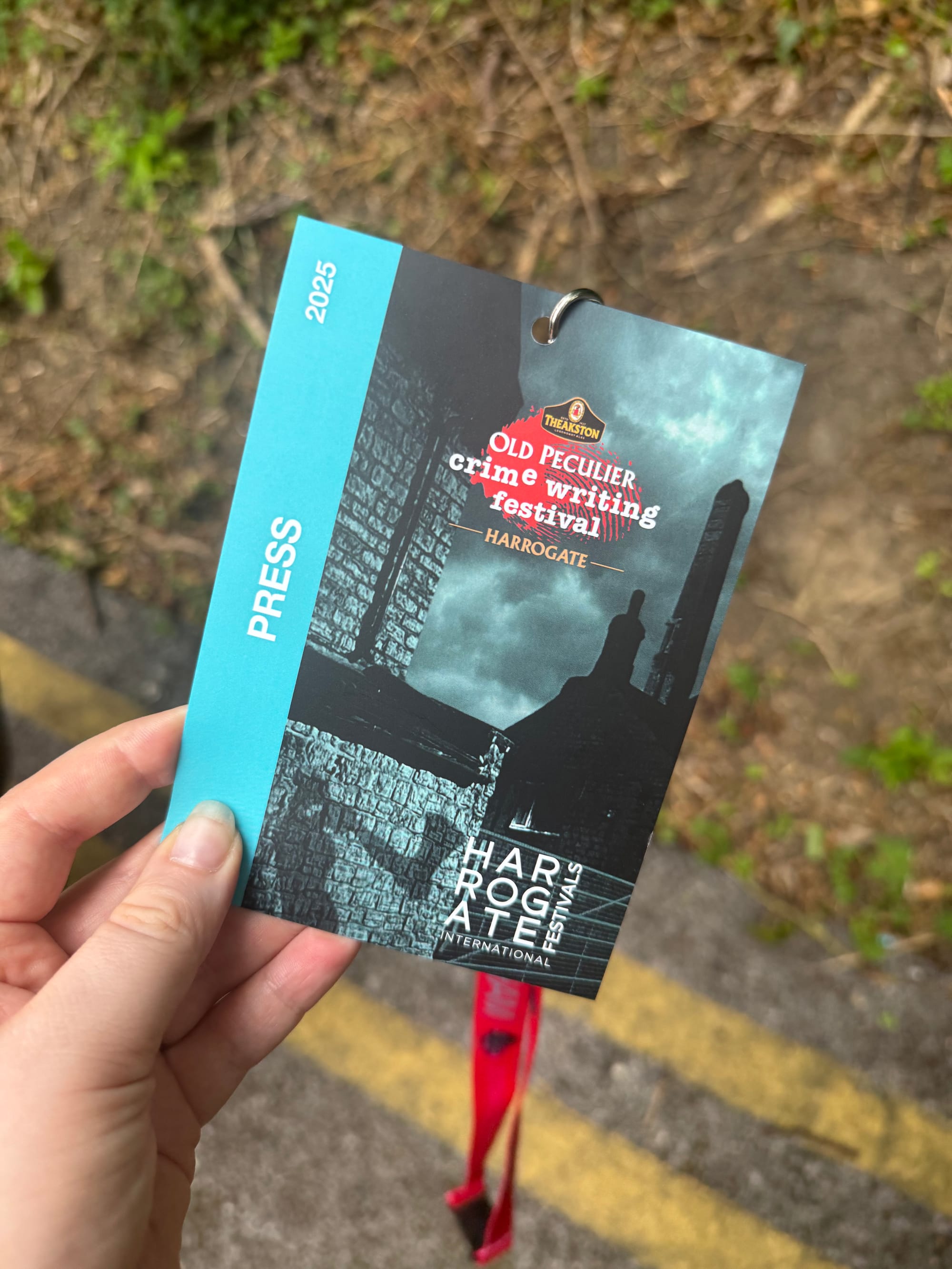 Close-up of a 2025 Theakston Old Peculier Crime Writing Festival press pass being held up by hand, with a red lanyard and a leafy outdoor background.
