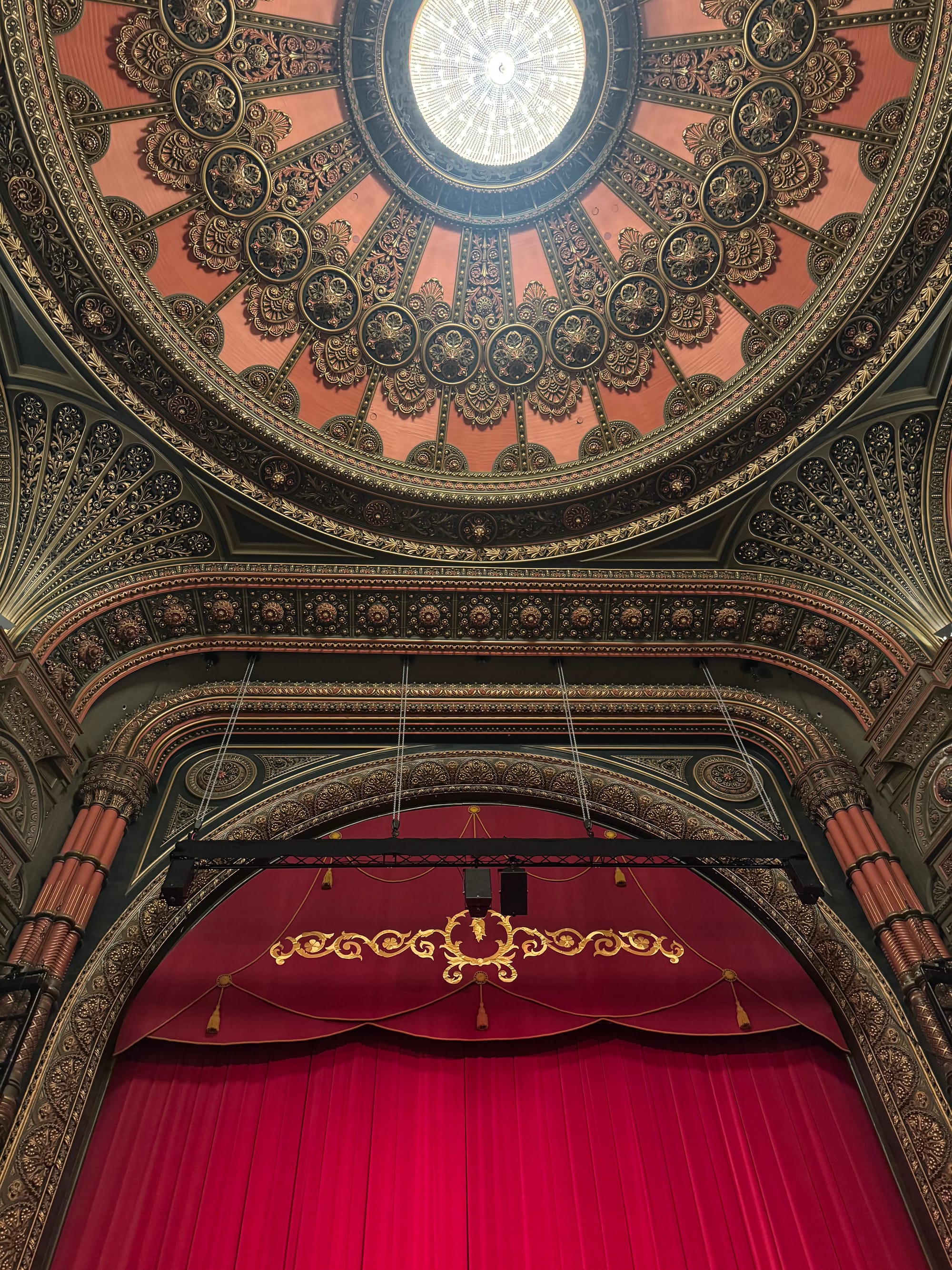 Ornate theater interior with a detailed domed ceiling and a luxurious red curtain featuring gold embroidery. Majestic and elegant atmosphere.