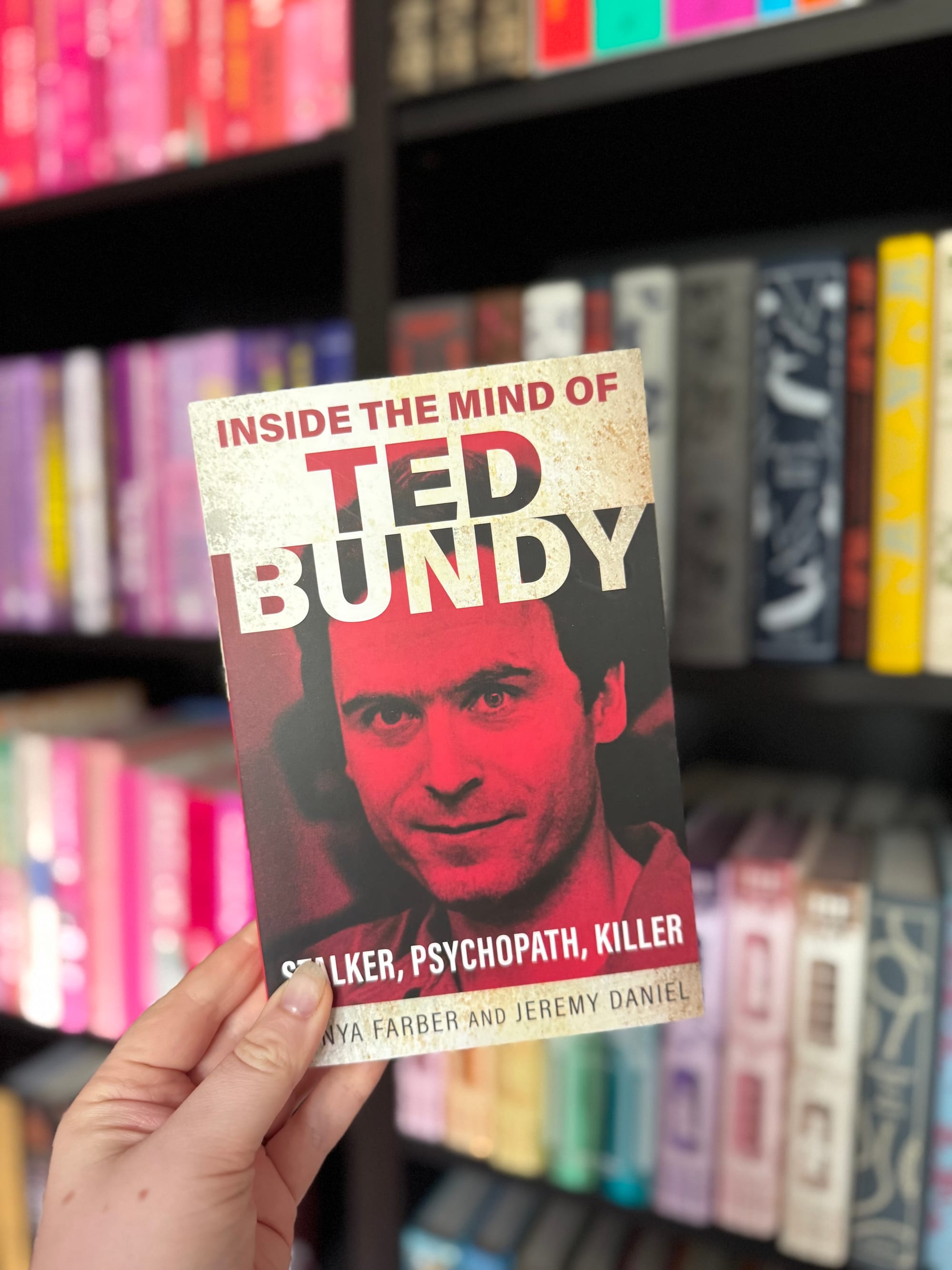 Hand holds a red-toned book titled "Inside the Mind of Ted Bundy" in a colorful library setting. Visible text includes "Stalker, Psychopath, Killer."