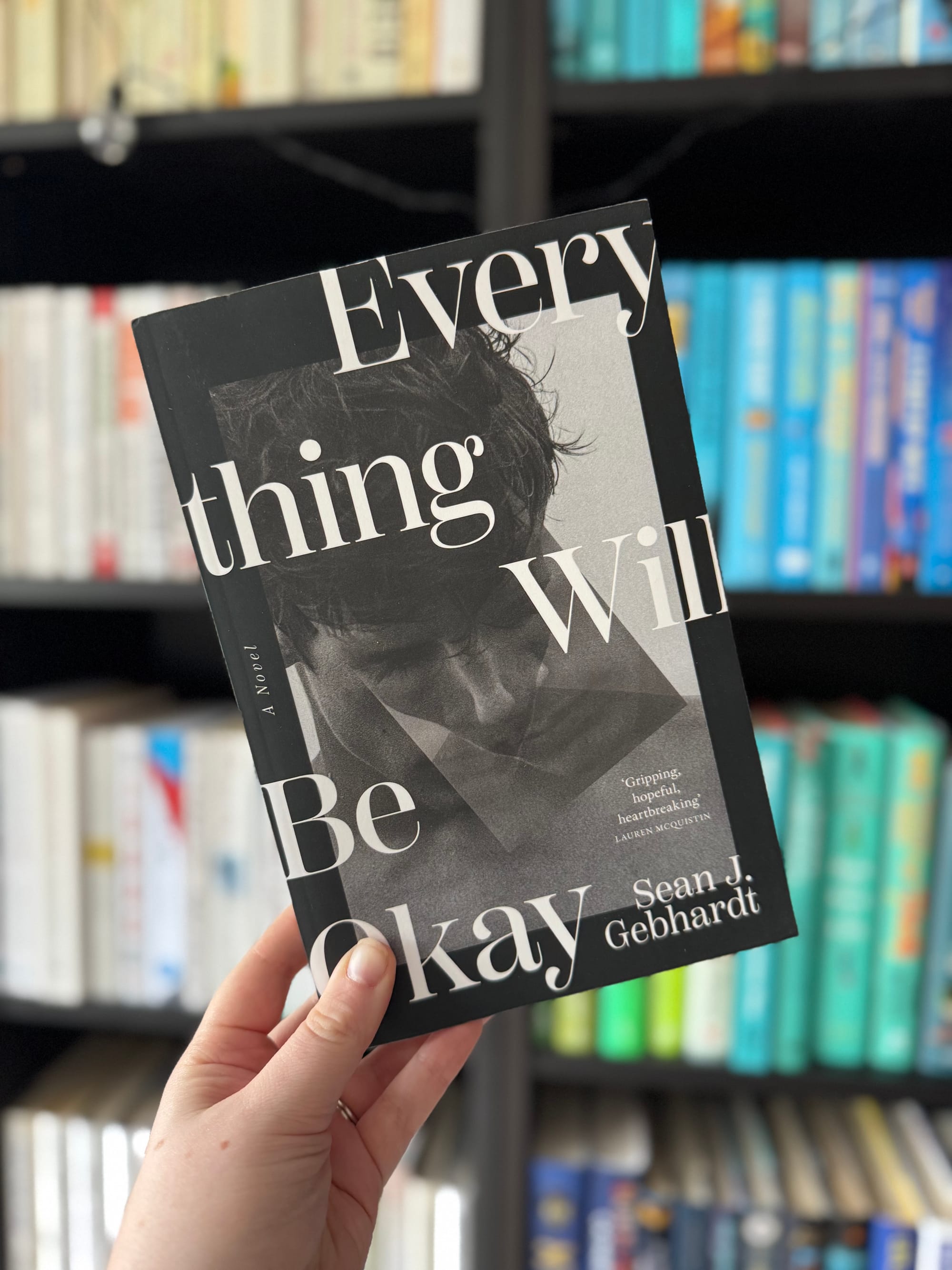 Hand holding a book titled "Everything Will Be Okay" by Sean J. Gebhardt in front of a bookshelf with colorful books. Mood: hopeful.