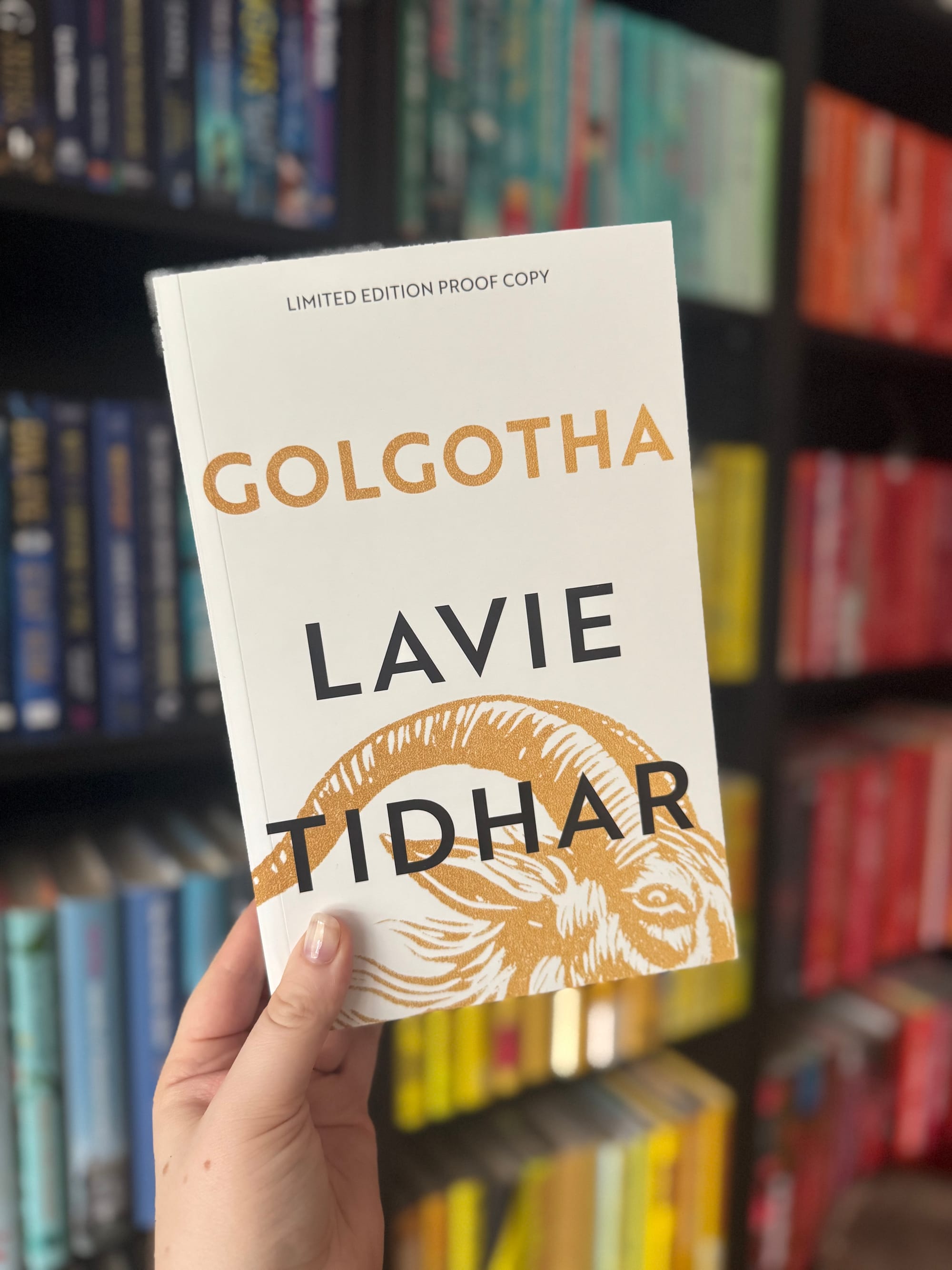 Hand holding a book titled "Golgotha" by Lavie Tidhar, with "Limited Edition Proof Copy" text. Background: colorful bookshelf, creating a vibrant mood.