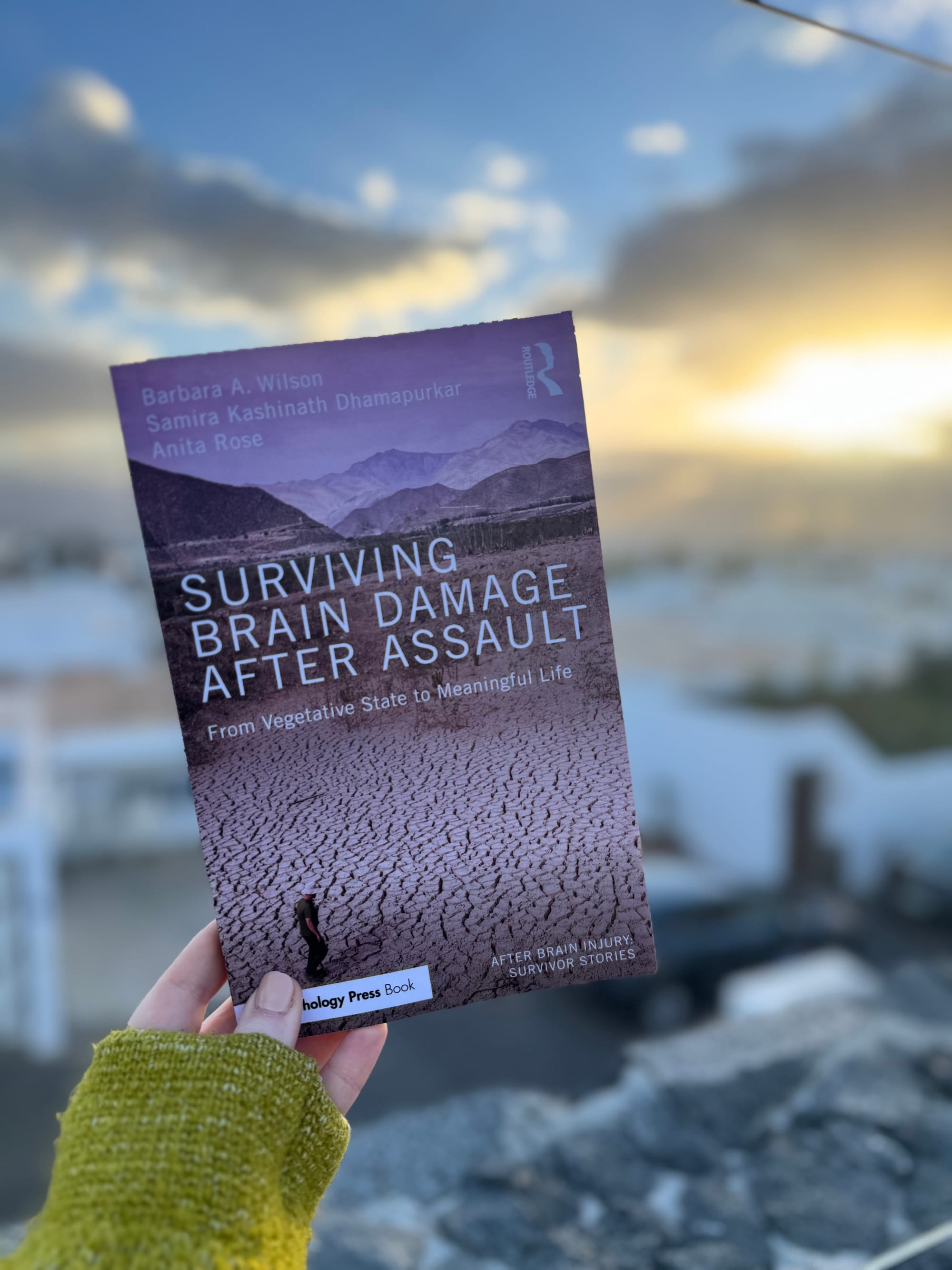 Person holding a book titled "Surviving Brain Damage After Assault" against a sunset sky. The cover shows a dry, cracked earth landscape.