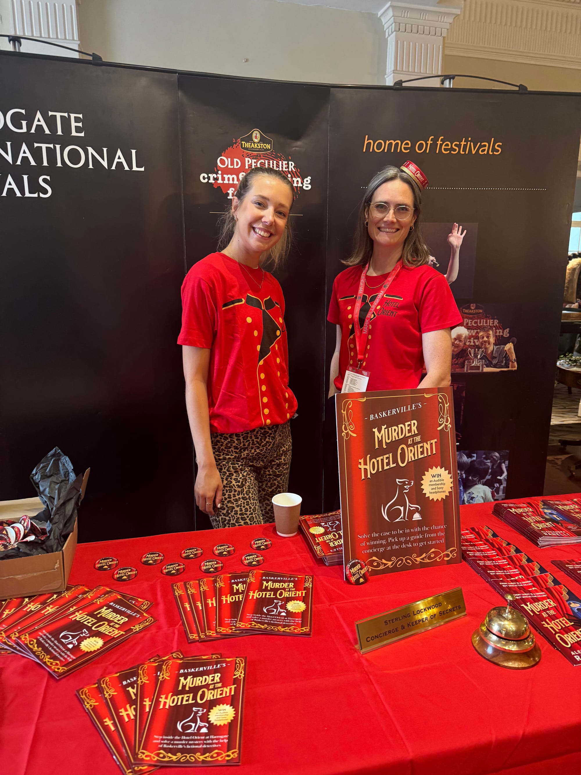 Two women in red shirts stand at a festival booth with "Murder at the Hotel Orient" books. Background text: "HARROGATE INTERNATIONAL FESTIVALS."