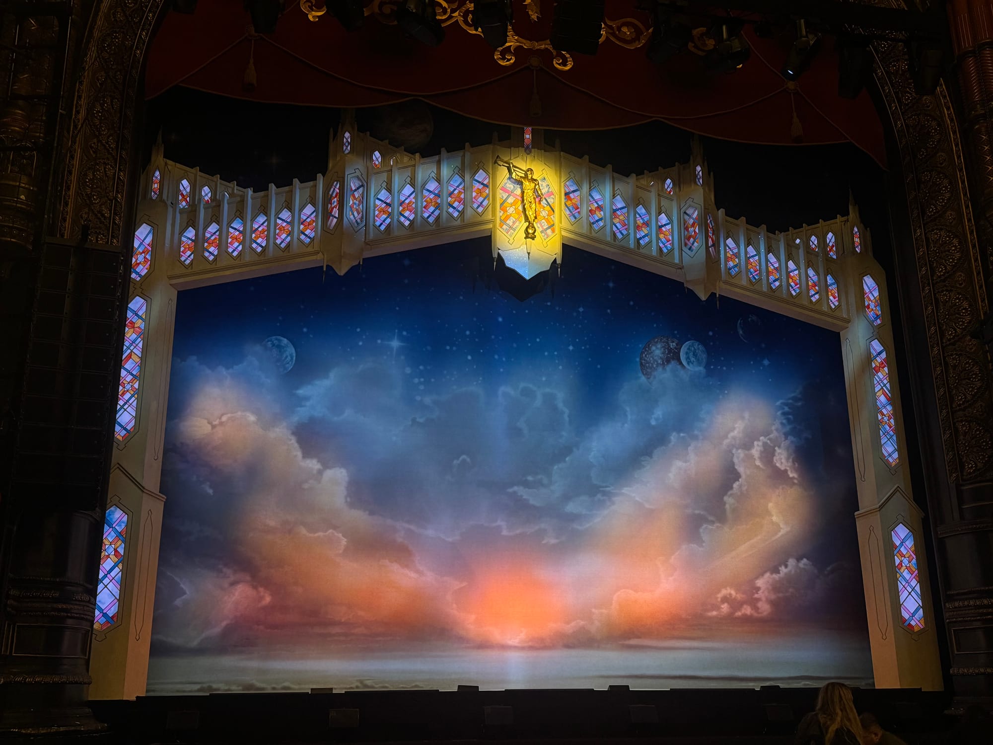 Stage set in a theater with vibrant clouds, planets, and a sunset backdrop. Gothic arch with stained glass and a glowing figure above.