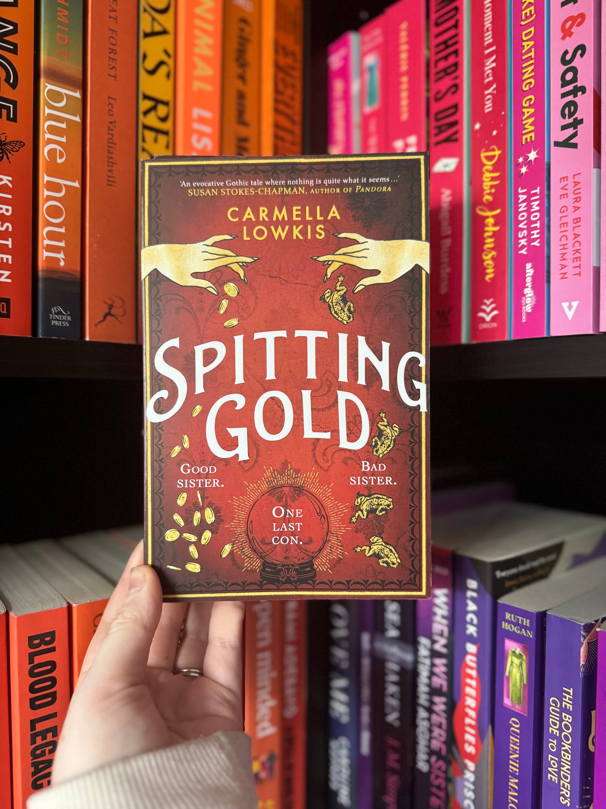 Hand holding the book "Spitting Gold" by Carmella Lowkis against a colorful bookshelf. The cover is red with coins and hands.