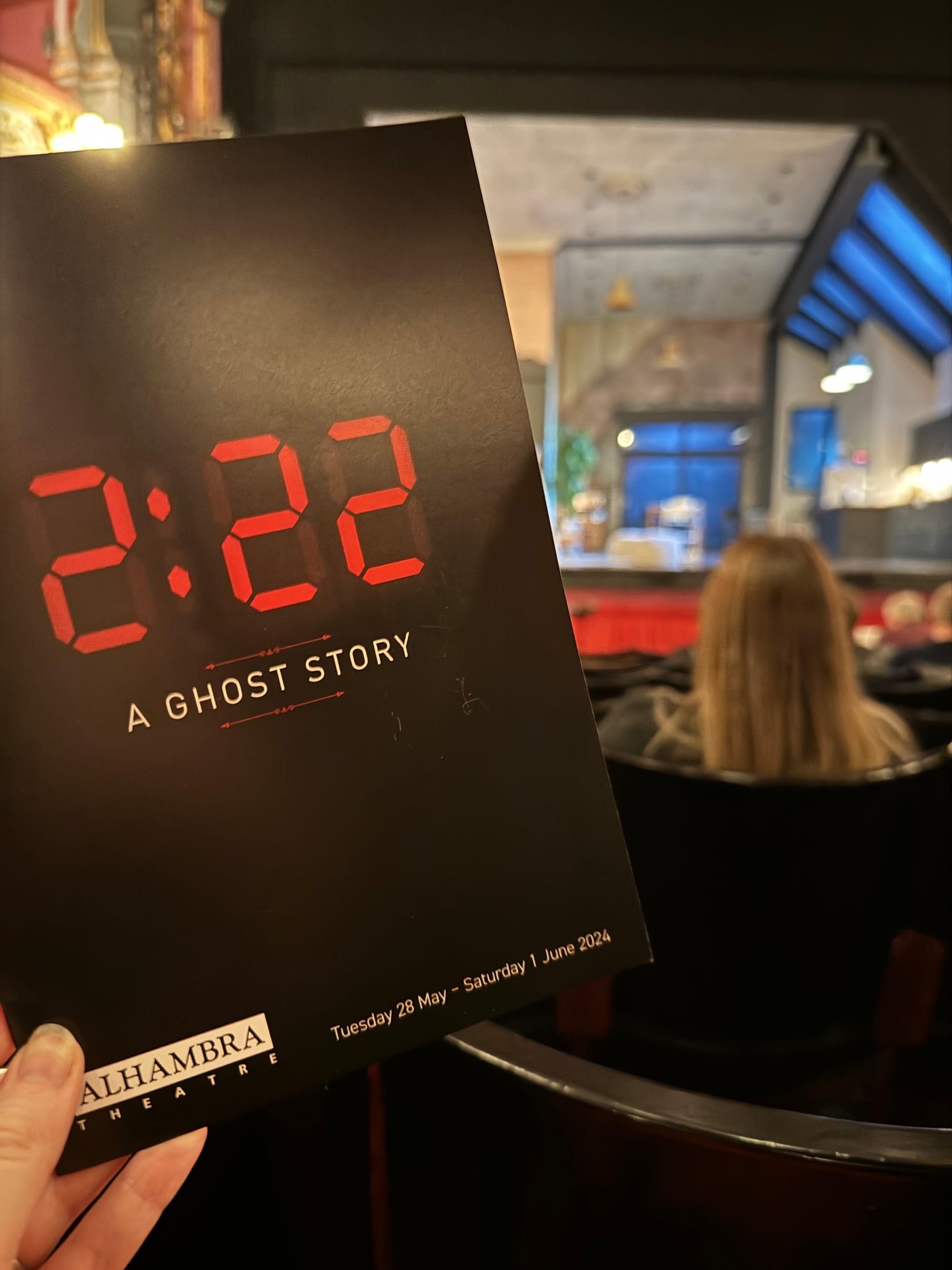 A theater attendee holds a brochure for "2:22 A Ghost Story" at Alhambra Theatre. Background shows a stage set for a play.