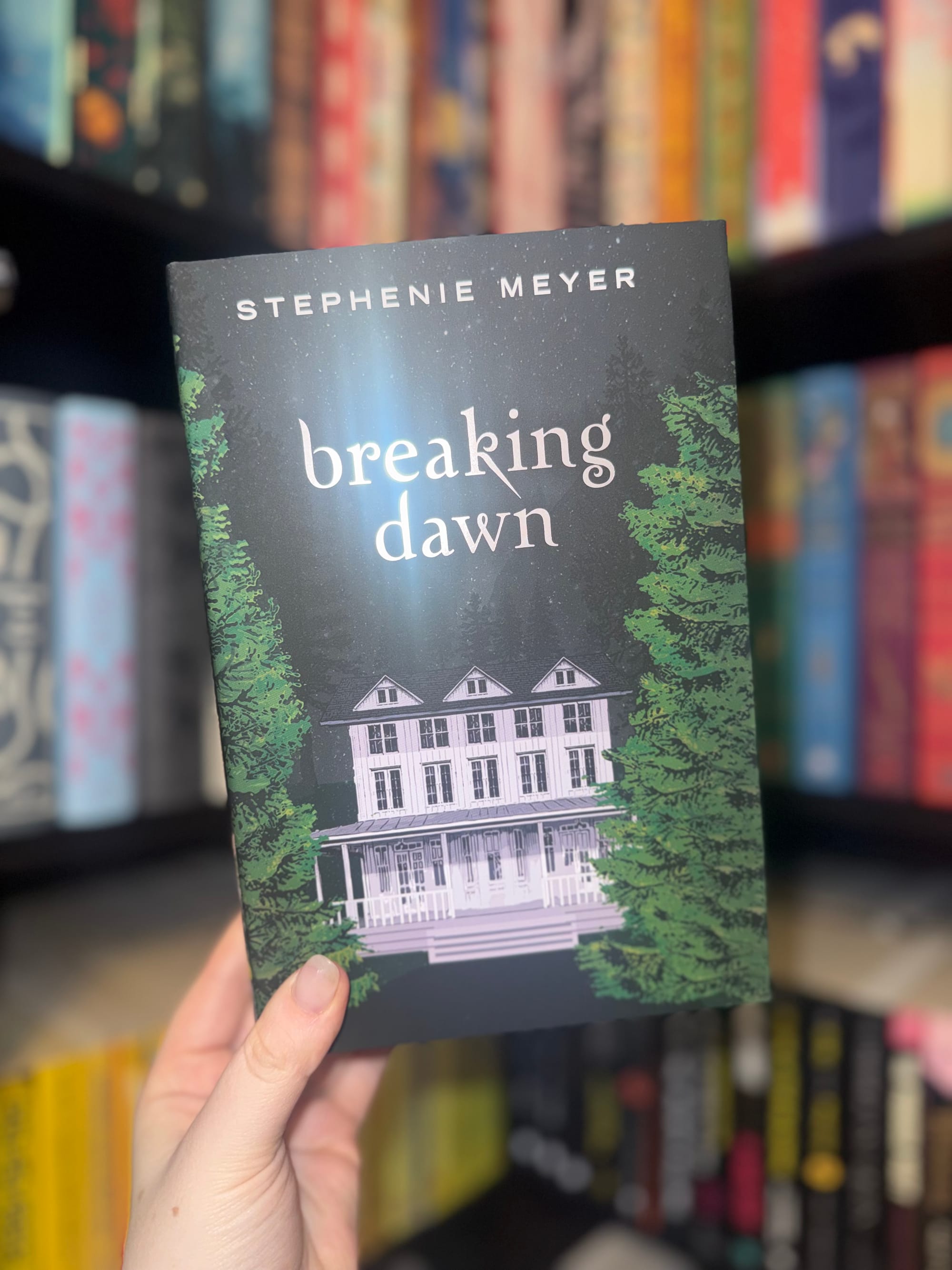 Hand holding "Breaking Dawn" by Stephenie Meyer, with a house and trees on the cover. Background shows colorful, blurred bookshelves.