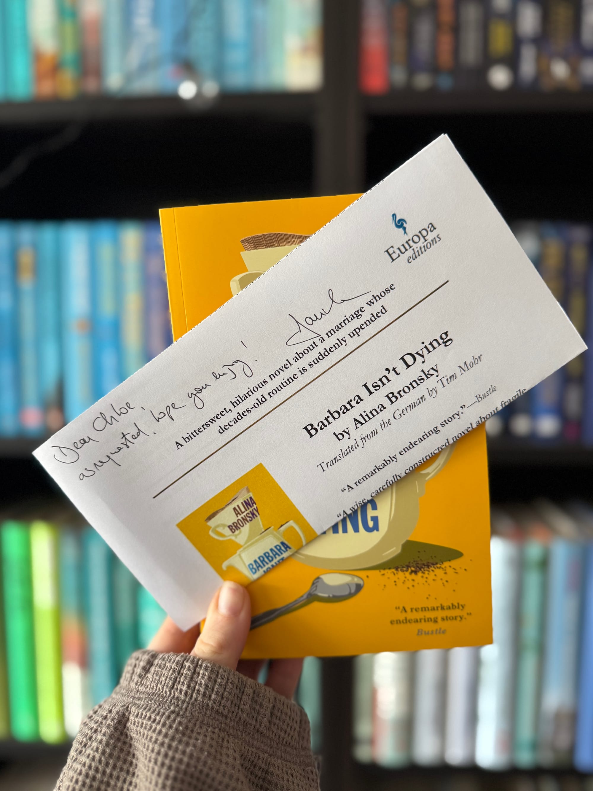Hand holding a yellow book titled "Barbara Isn't Dying" and a note with "Dear Chloe, enjoy!" in front of colorful bookshelves.
