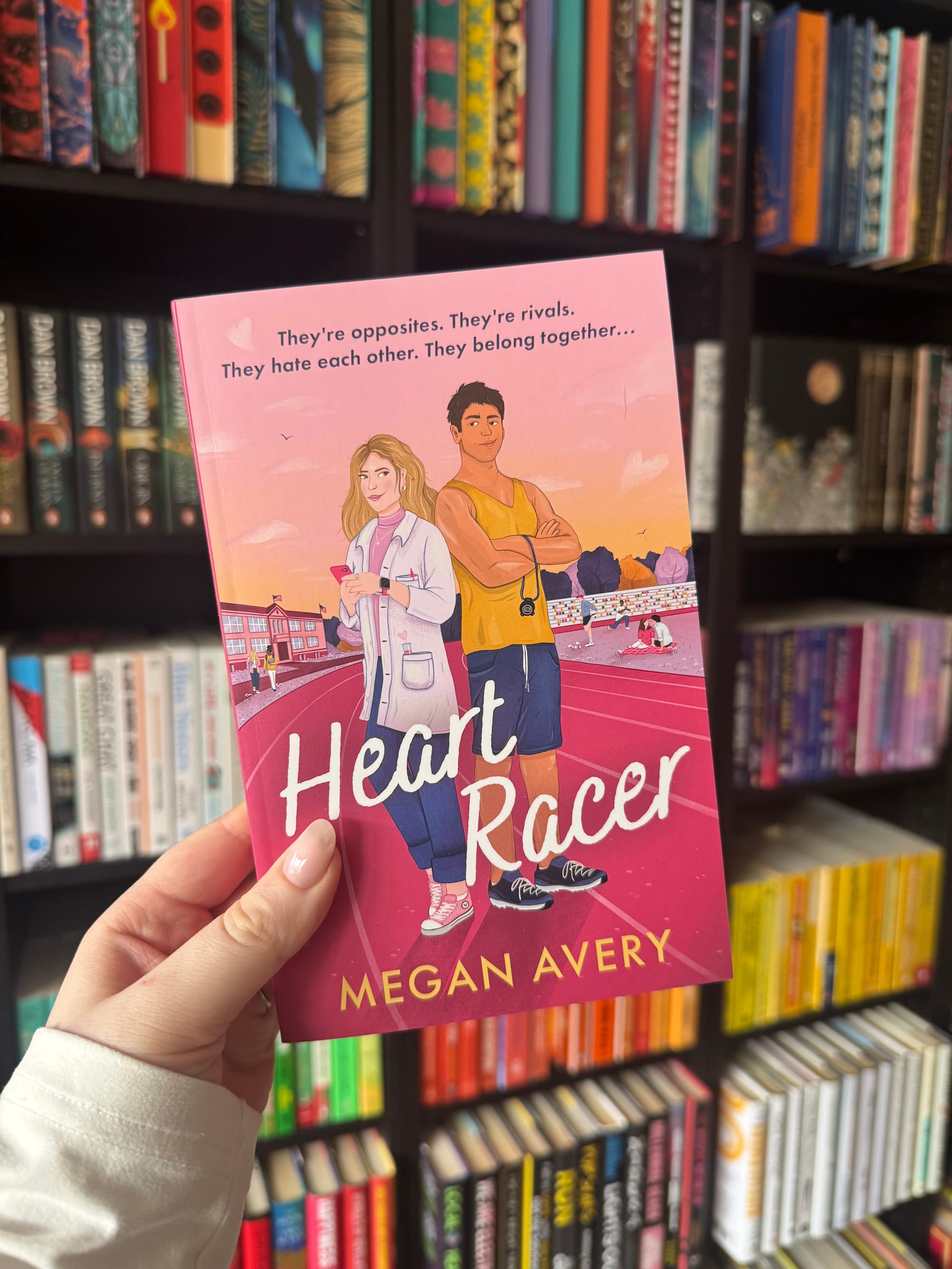 Person holding a pink book titled "Heart Racer" by Megan Avery, featuring illustrated characters. Bookshelves with colorful books in background.