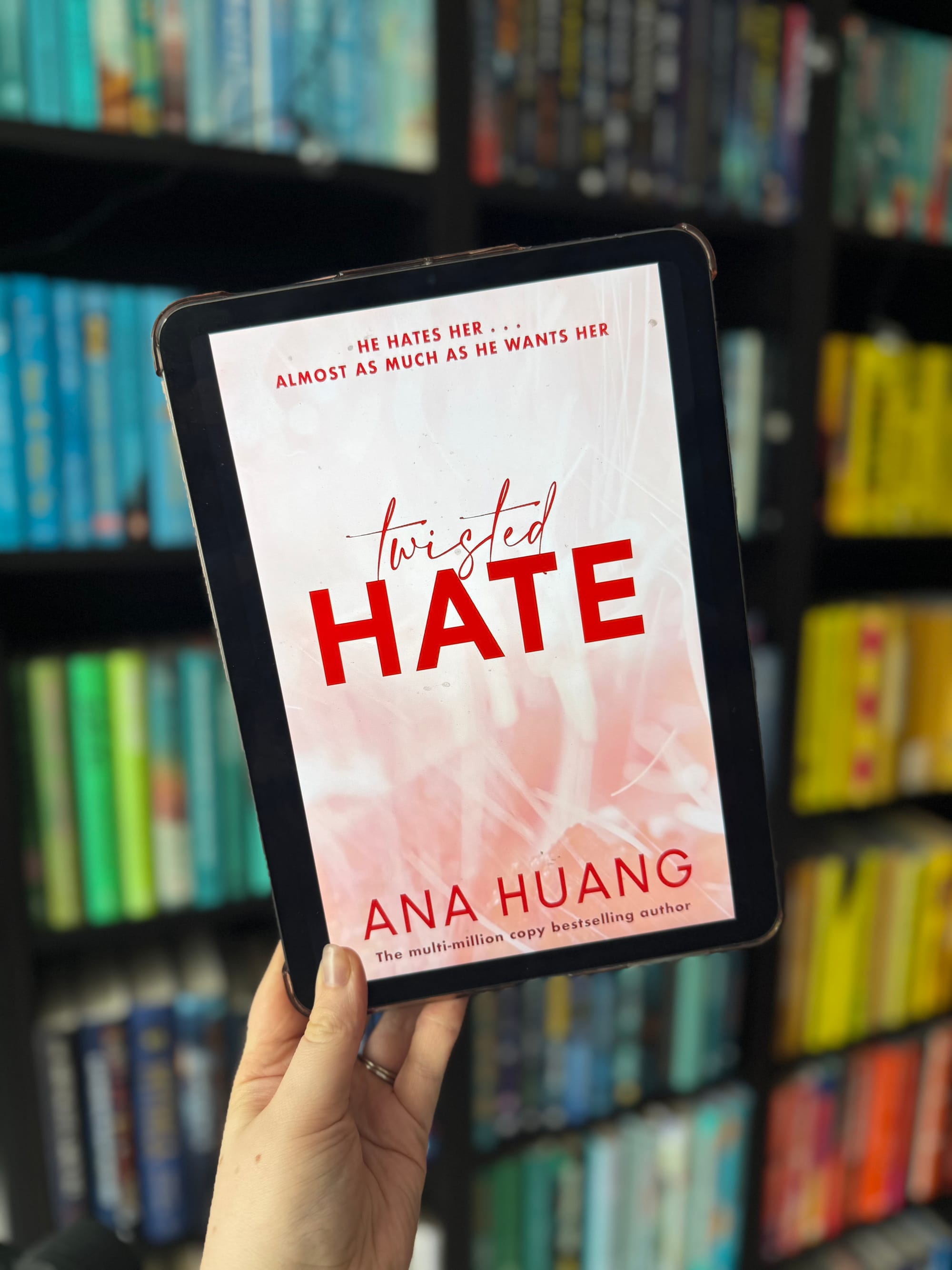 A hand holds a tablet showing the book "Twisted Hate" by Ana Huang. Background has colorful bookshelves creating a vibrant setting.