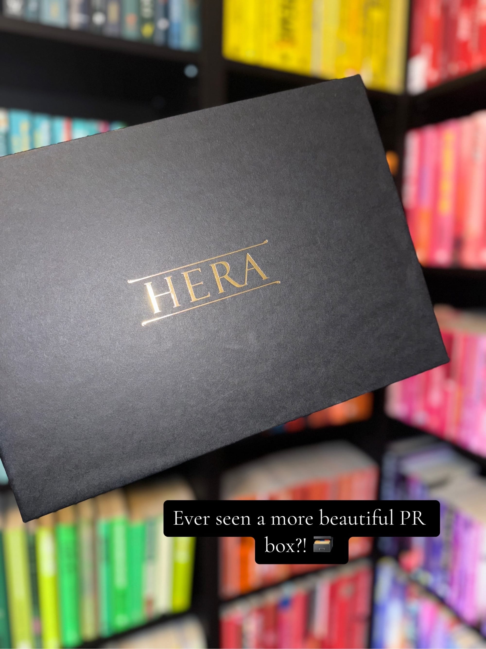 Black PR box with gold "HERA" text, held in front of colorful bookshelves. Text overlay: "Ever seen a more beautiful PR box?!" π