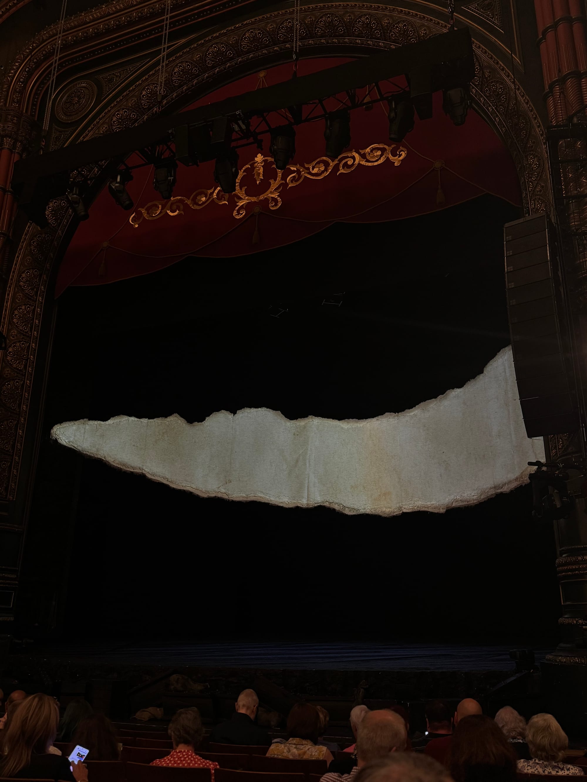 The stage of a theatre before a performance of War Horse. A large torn parchment-style screen hangs at the centre, lit against the dark stage. Audience members are seated in the foreground, waiting for the show to begin.