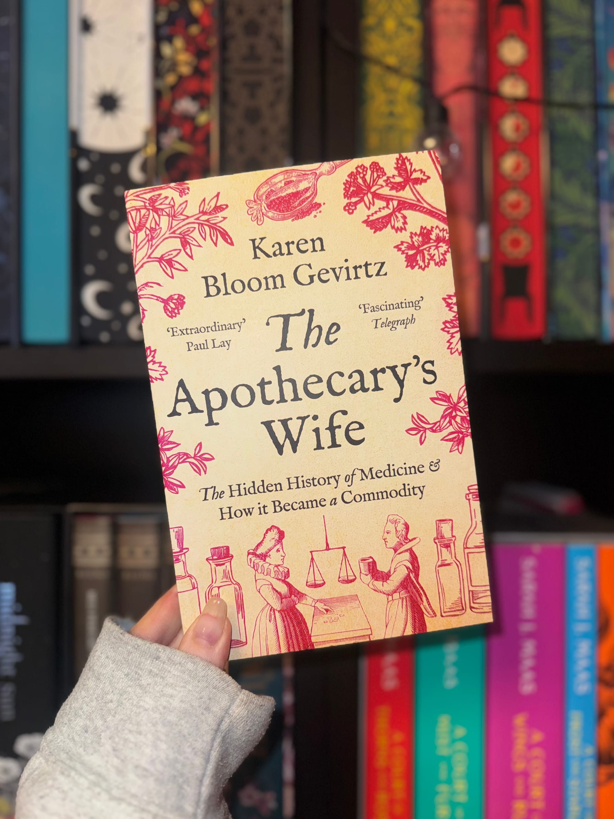 A hand holds "The Apothecary's Wife" by Karen Bloom Gevirtz in front of colorful books on a shelf. The cover features vintage illustrations.