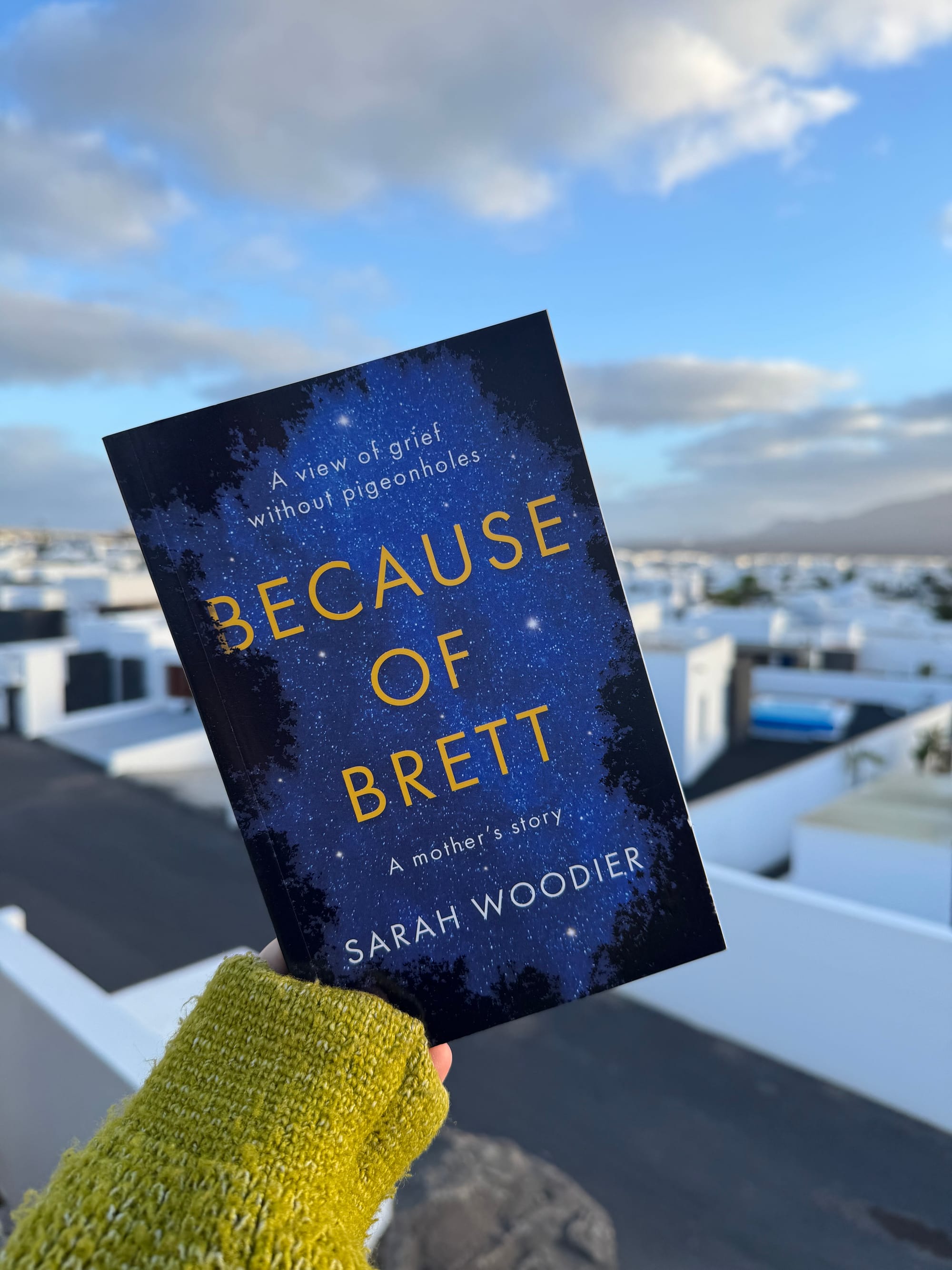 Person holding "Because of Brett" book with a starry cover against a cloudy sky and white buildings background, wearing a green knit cardigan.