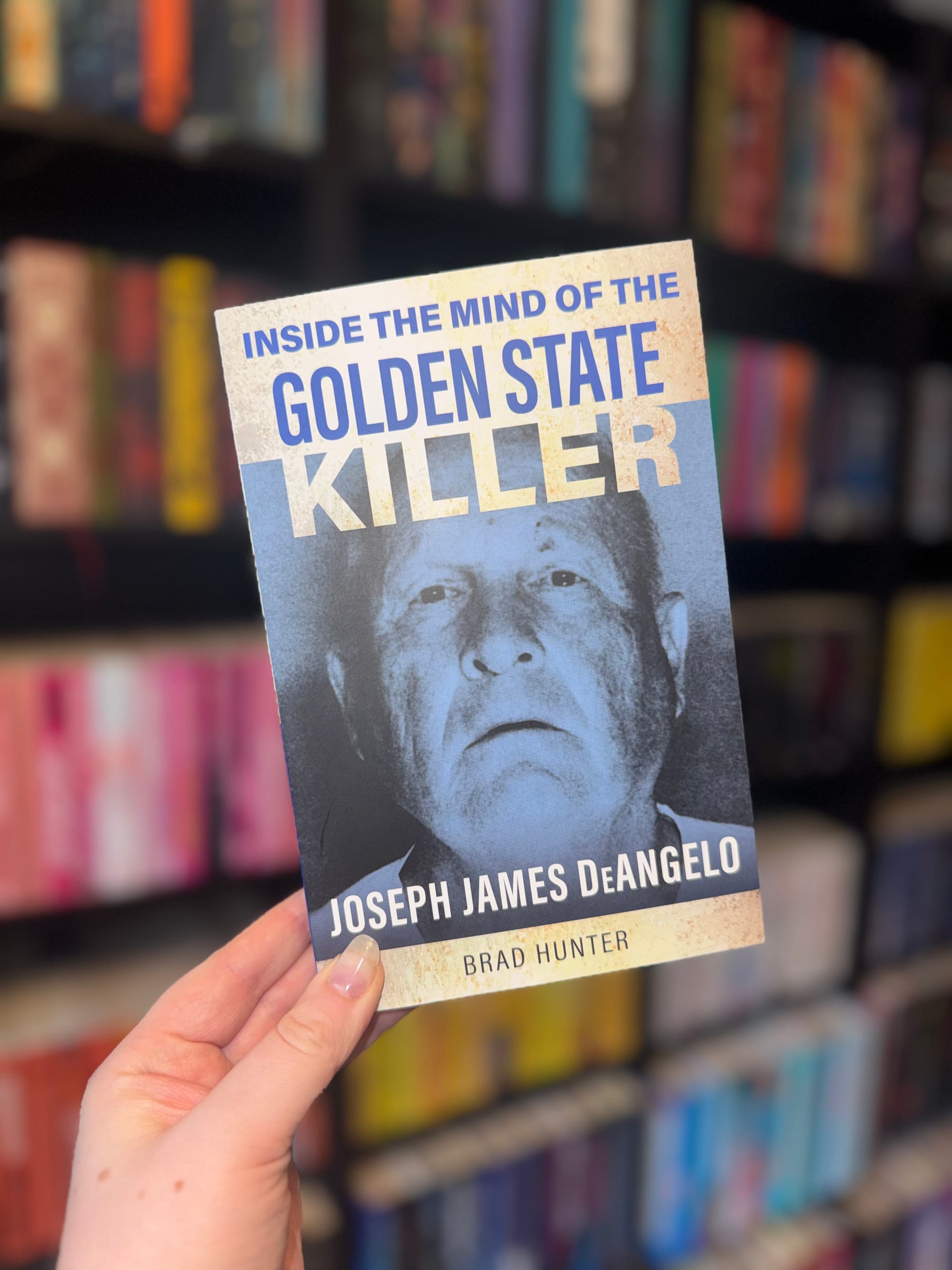 Hand holding a book titled "Inside the Mind of the Golden State Killer" by Brad Hunter. Blurred bookstore shelves in the background.