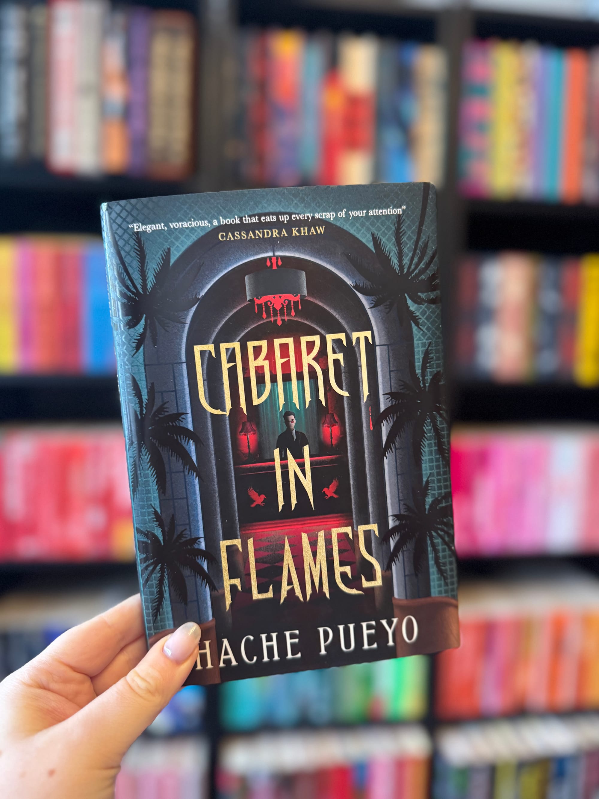 Hand holds "Cabaret in Flames" book by Hache Pueyo, against a blurred colorful bookshelf. Cover shows palm trees, red lights, dramatic.