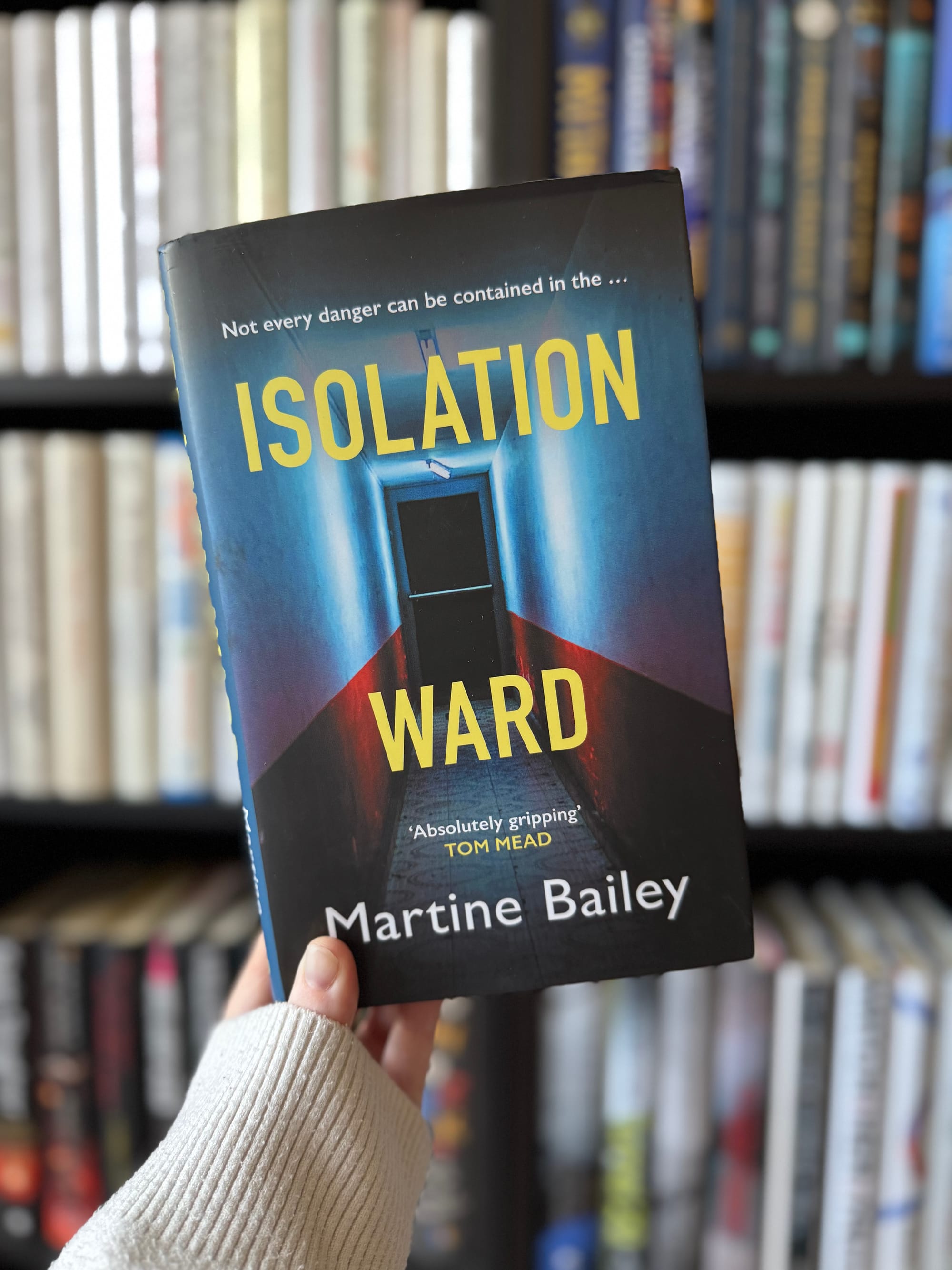 Person holds the book "Isolation Ward" by Martine Bailey. Cover shows a dark hallway. Bookshelf in blurred background. Mood is suspenseful.