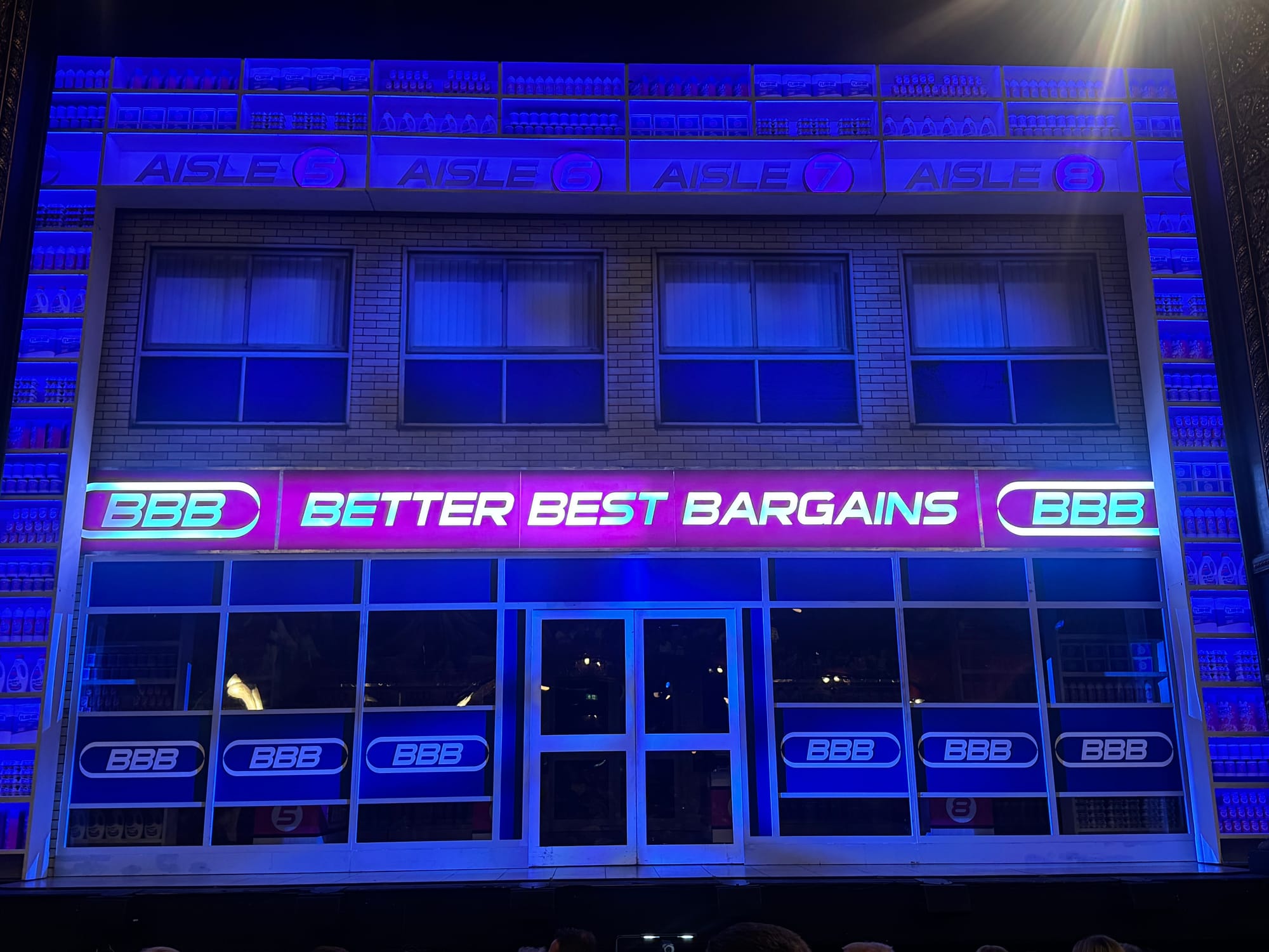 Brightly lit store facade labeled "BBB BETTER BEST BARGAINS" at night, with neon signs and shelves displayed above. Blue and purple hues.