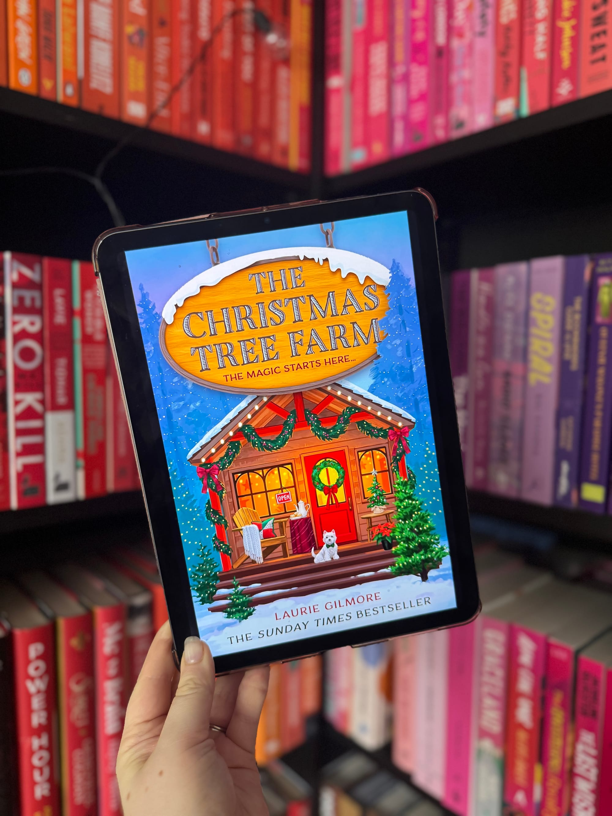 Hand holding a tablet displaying "The Christmas Tree Farm" book cover with a festive cabin. Colorful bookshelves in the background.