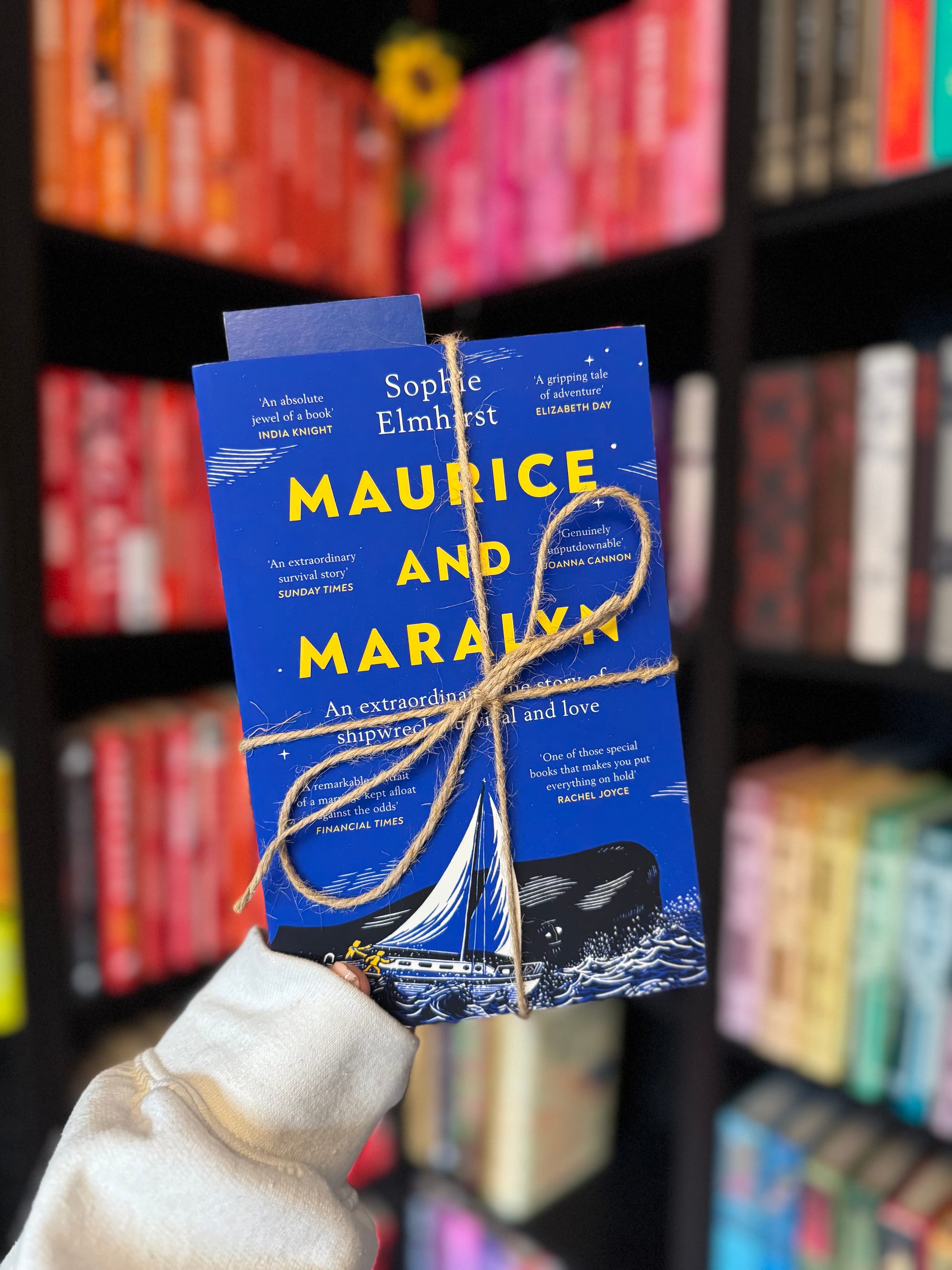 A hand holds a blue book titled "Maurice and Maralyn" by Sophie Elmhirst, tied with twine. Colorful bookshelves in the blurred background.