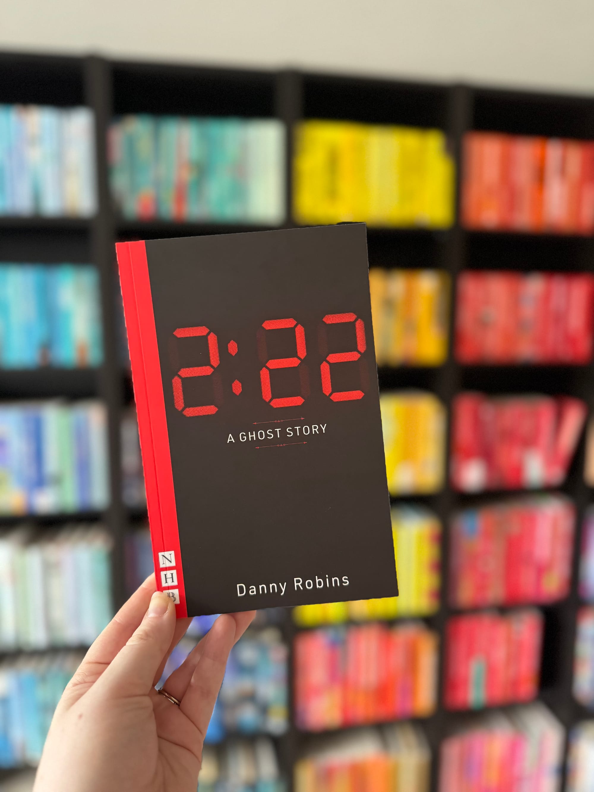 Hand holding "2:22 A Ghost Story" by Danny Robins with colorful bookshelf in the background. The book cover is black with red text.