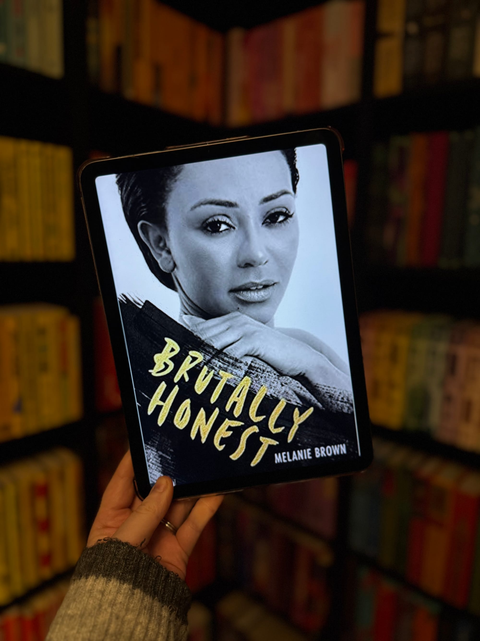 A hand holds a tablet displaying "Brutally Honest" by Melanie Brown. The background shows colorful, blurred bookshelves.