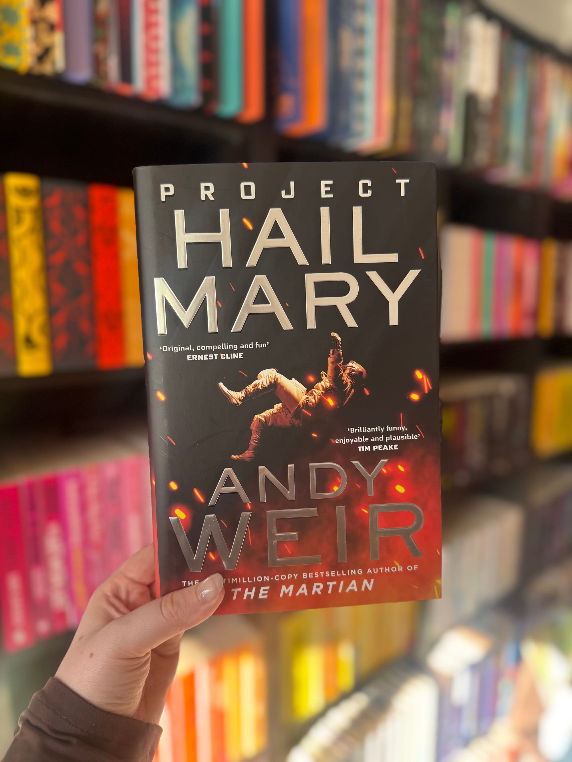 A hand holds the book "Project Hail Mary" by Andy Weir in a bookstore. The cover shows an astronaut in space with a colorful book display behind.