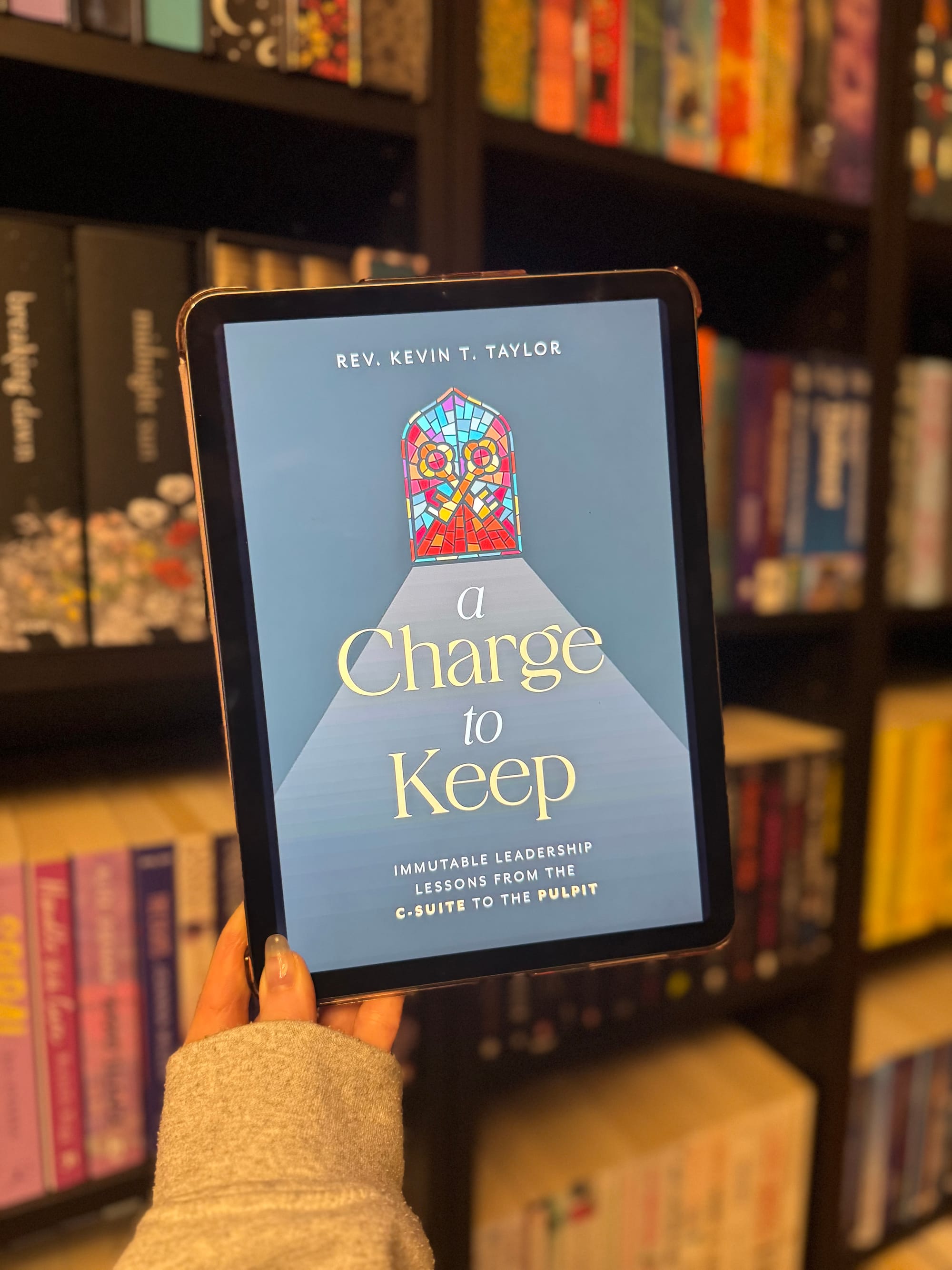 Person holding a tablet displaying Rev. Kevin T. Taylor's book "A Charge to Keep" in front of a colorful bookshelf.