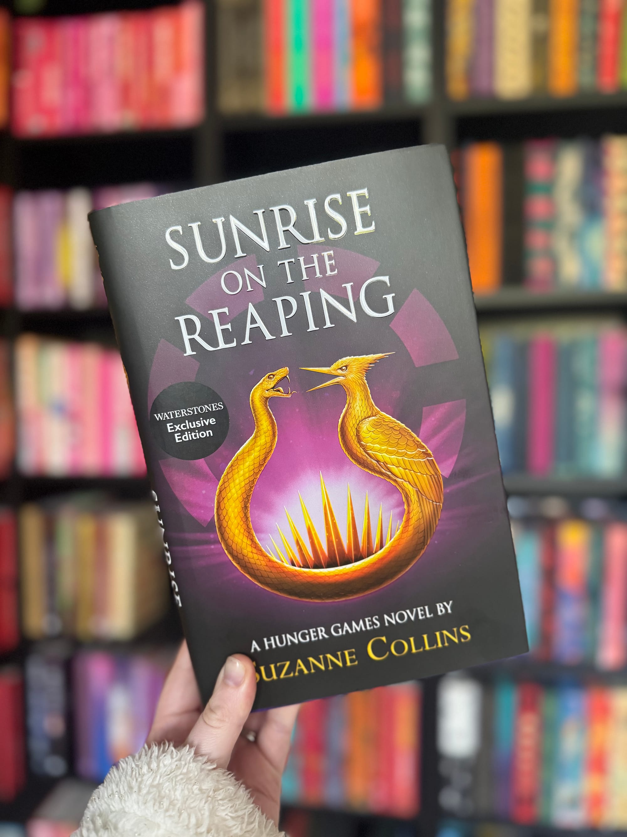 Hand holding "Sunrise on the Reaping" book with golden bird logo against a colorful bookshelf backdrop. Text: "Waterstones Exclusive Edition".